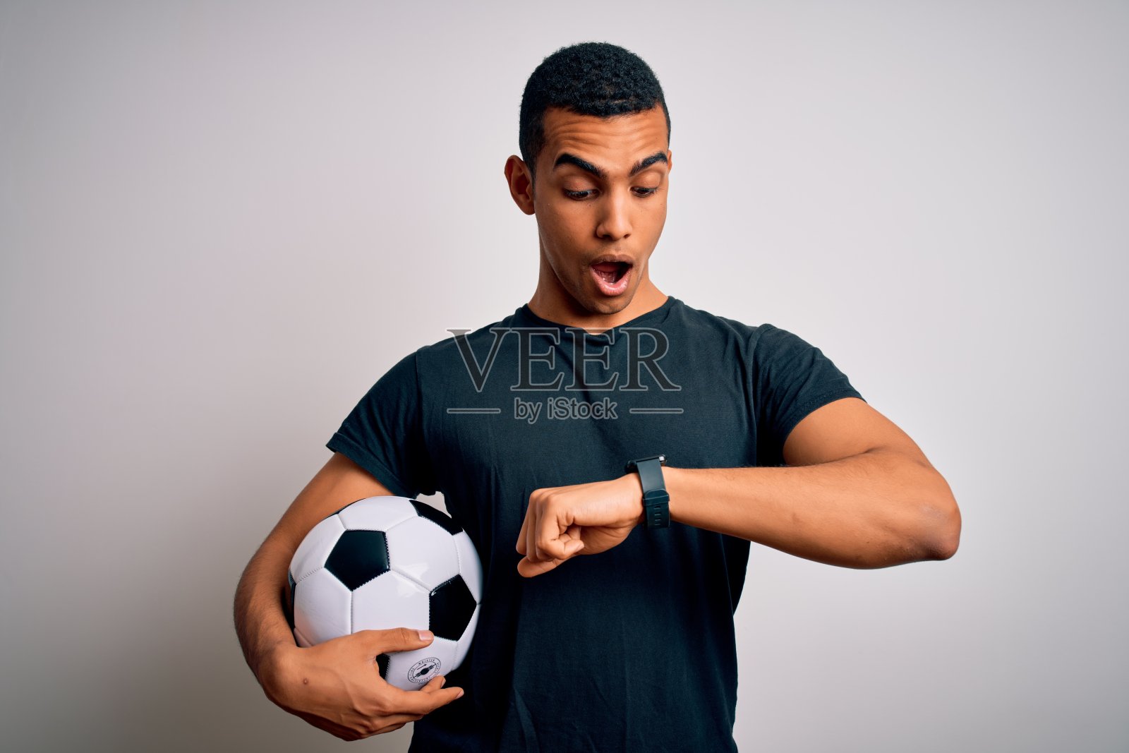 英俊的非裔美国人正在踢足球，手里拿着足球越过白色的背景看着手表时间着急，怕迟到照片摄影图片
