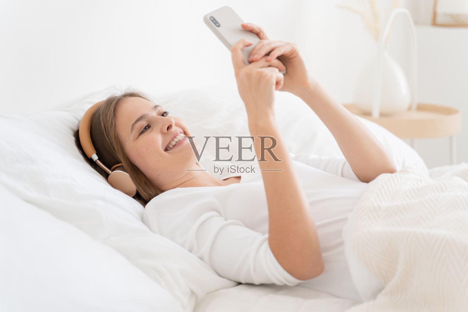 年轻女子躺在白色的床上，穿着上衣，一边玩一边通过无线耳机听她的朋友，享受智能手机上的视频通话照片摄影图片