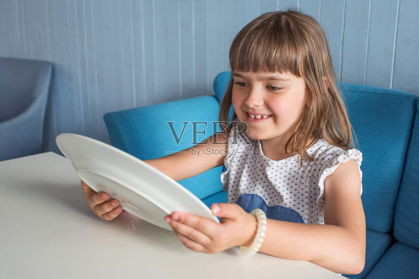 小女孩坐在桌旁，手里拿着一个空盘子。一个孩子微笑着看着一个空盘子。照片摄影图片