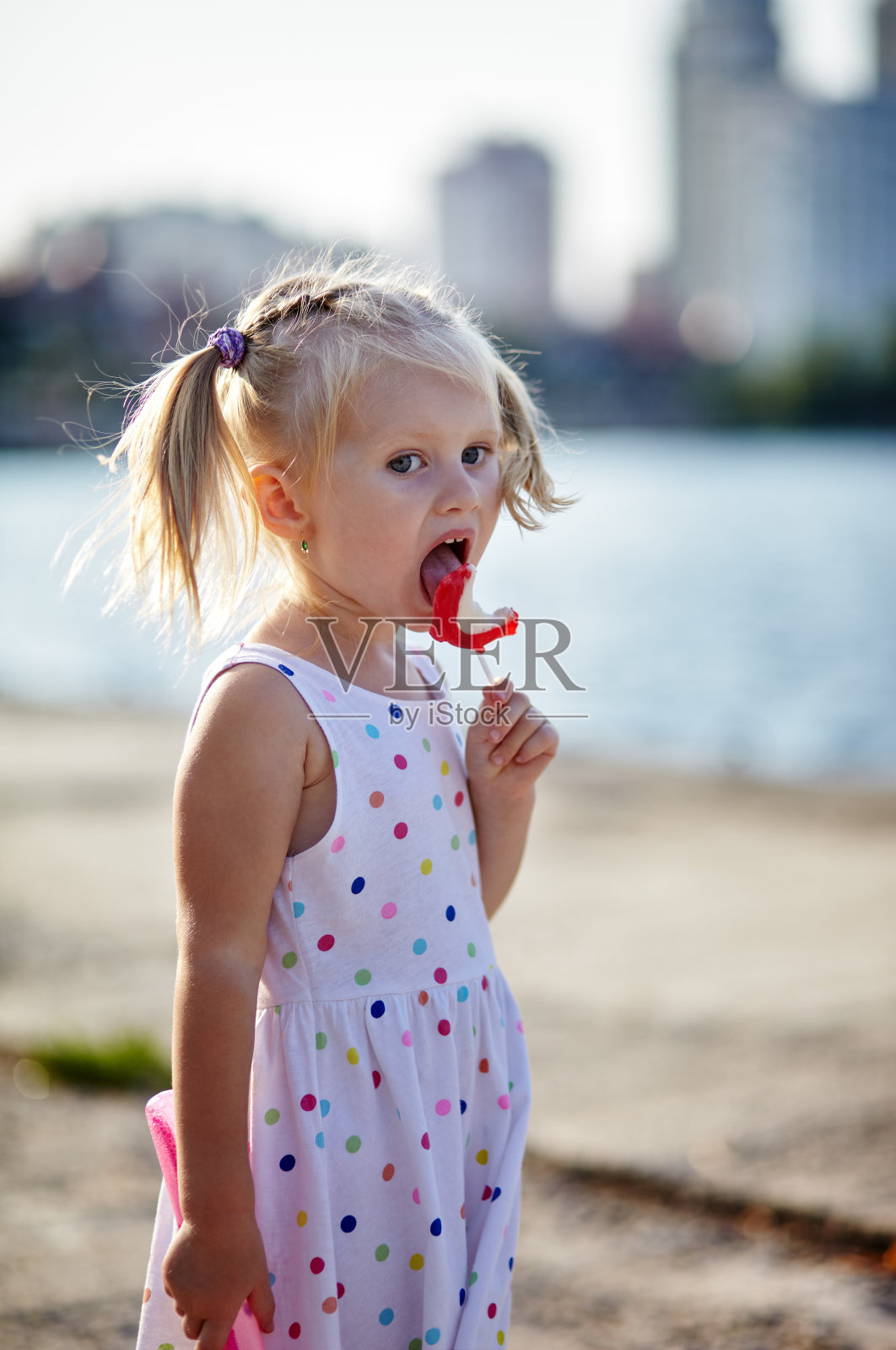可爱的孩子穿着白色连衣裙在户外摆姿势。女孩很开心。小女孩在公园里吃棒棒糖。暑假或春假照片摄影图片