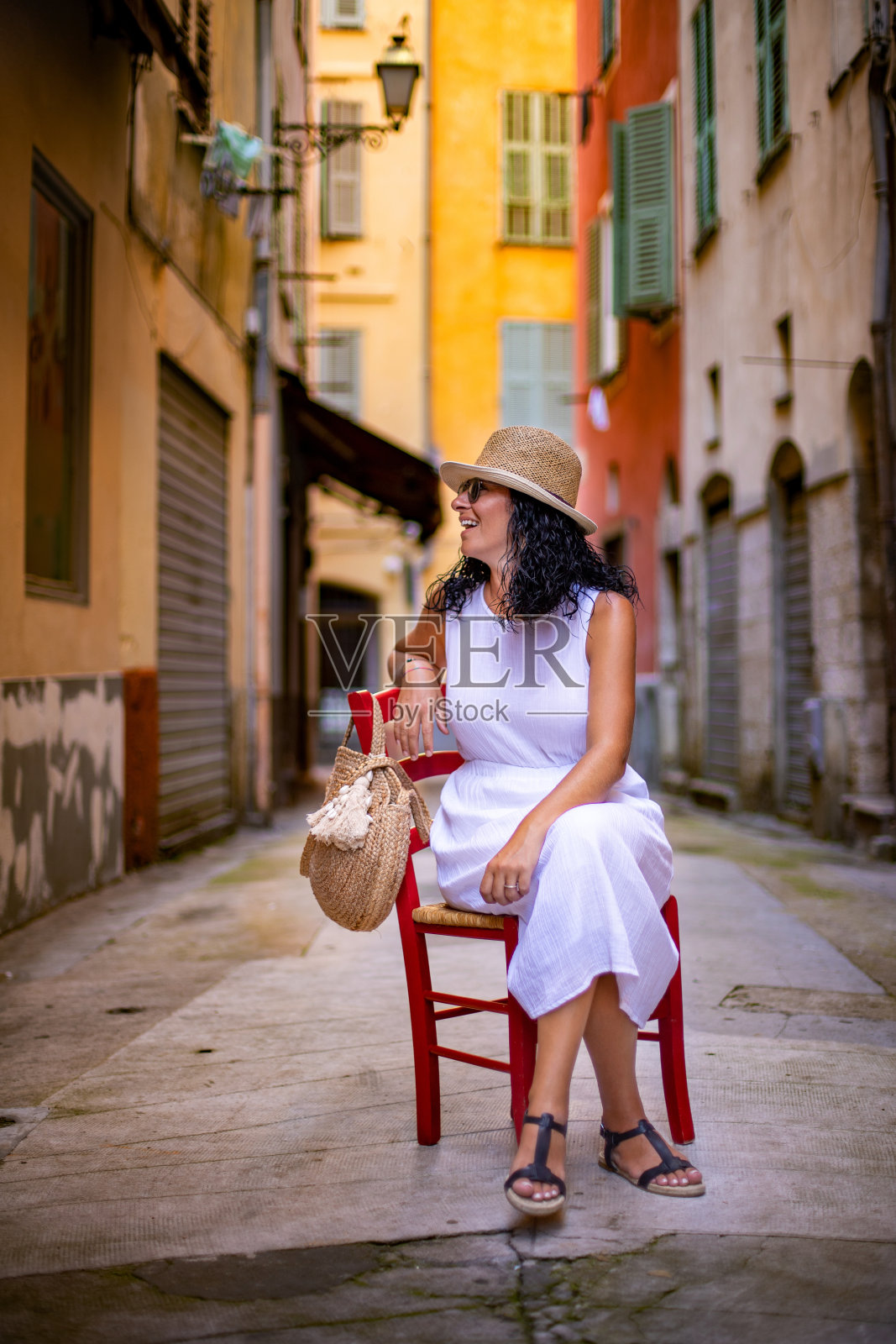 一位美丽的女人坐在法国尼斯五颜六色的街道上的木椅上。照片摄影图片