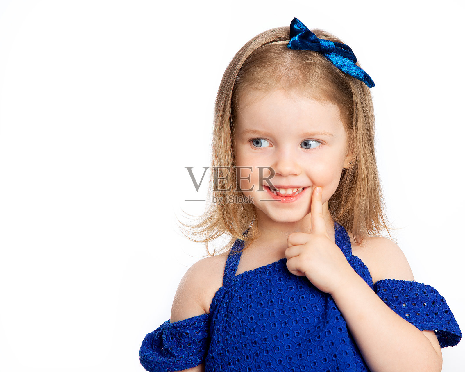 一个美丽的蓝眼睛小女孩看起来狡猾神秘，微笑愉快，并把手指放在她的脸颊。照片摄影图片