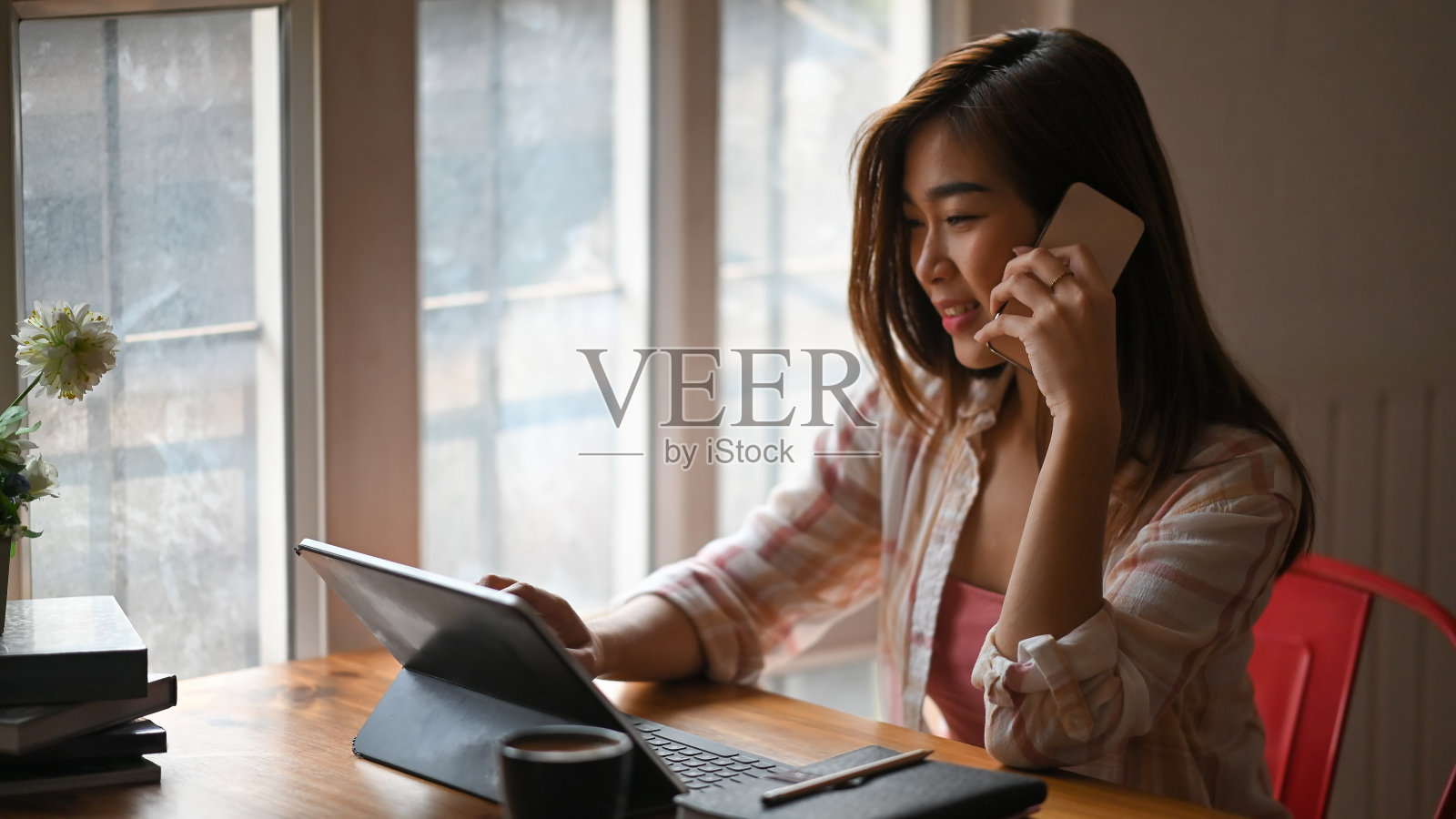 美丽的女人一边用平板电脑一边用手机聊天，坐在木制的办公桌前，舒适的客厅作为背景。照片摄影图片