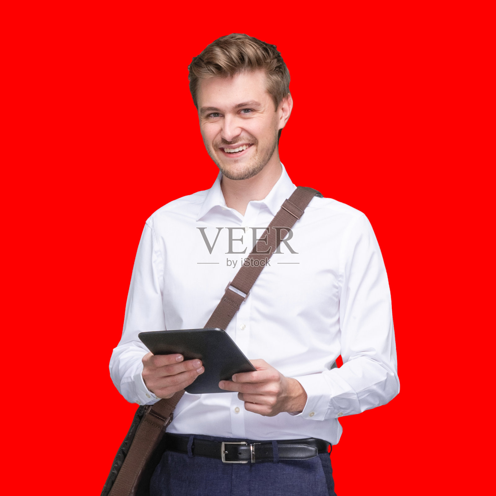 白人年轻男性经理站在有色背景前，戴着邮差包，拿着包，使用数码平板电脑照片摄影图片