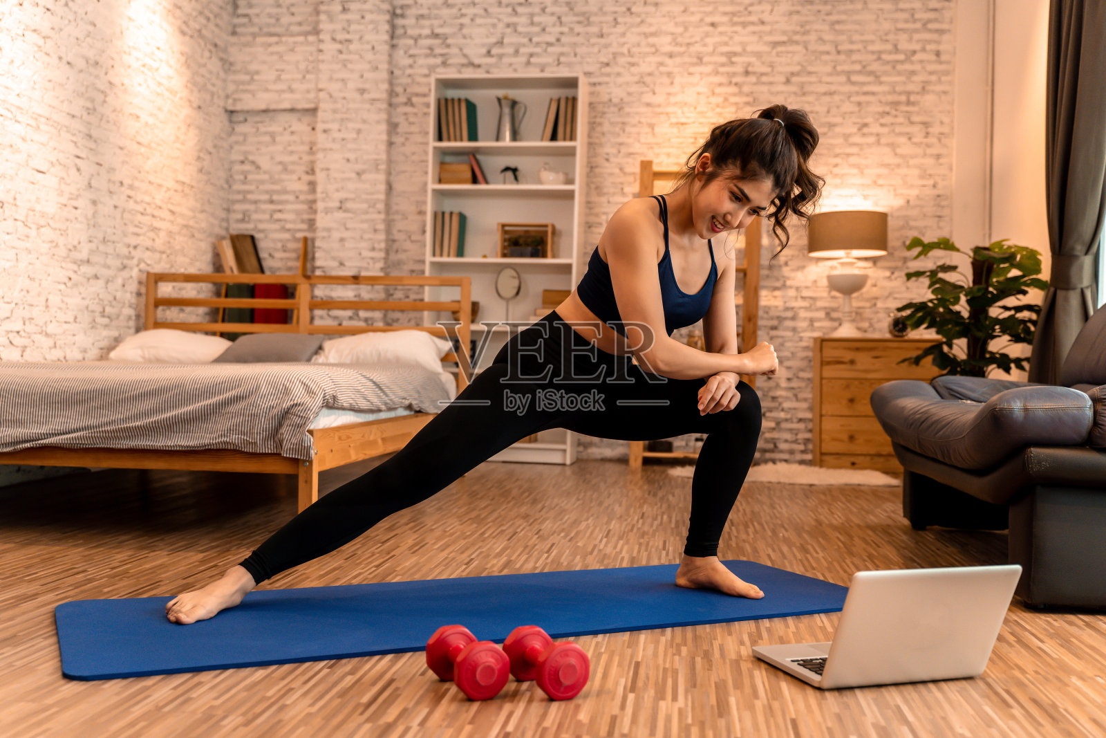 20多岁的年轻亚洲女子，穿着运动服，一边做伸展运动，一边在电脑上看瑜伽课。健康的女孩在客厅锻炼，背景是沙发。照片摄影图片
