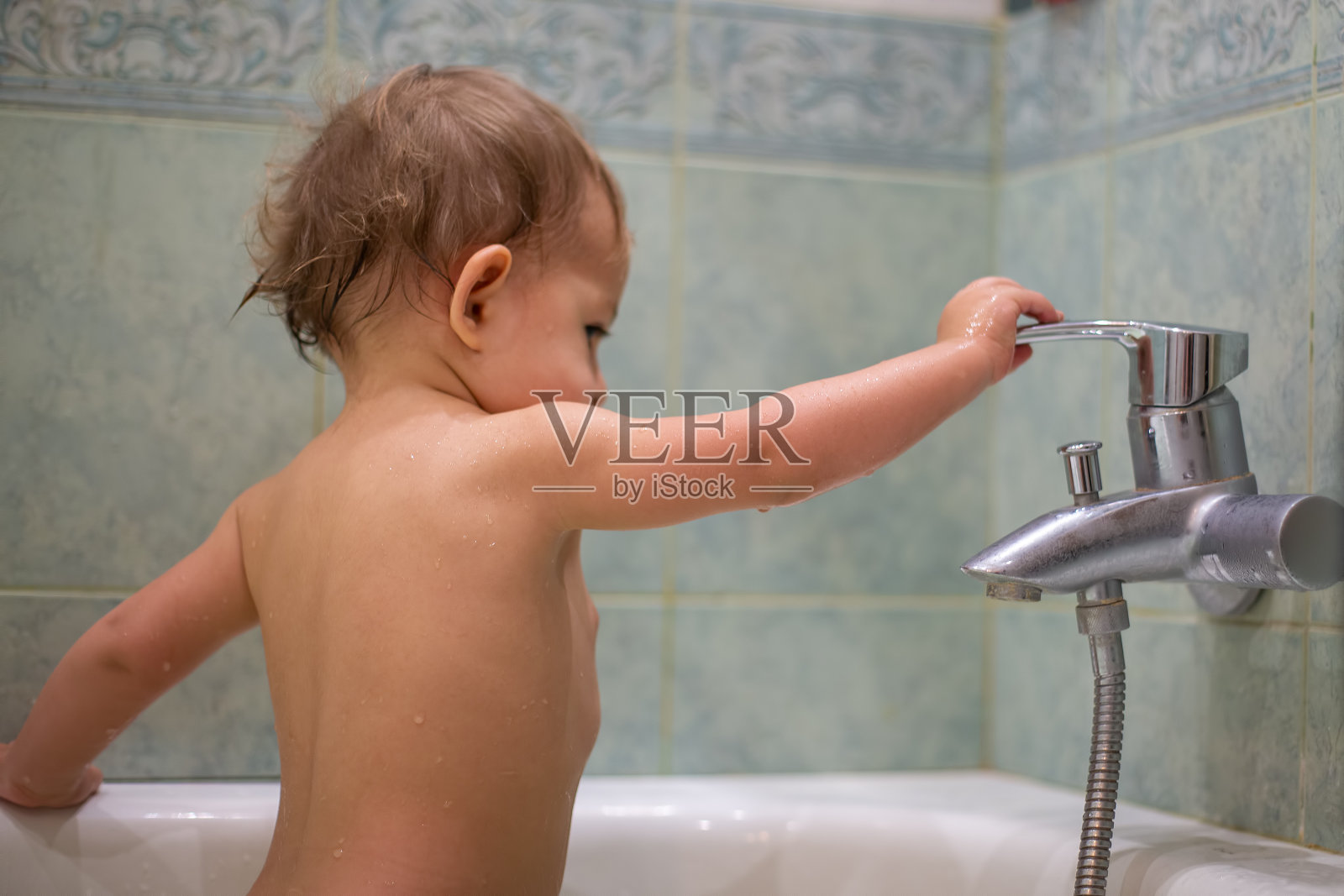 一个可爱的小孩，在洗澡的时候，玩着水龙头，开关它。以模糊的绿色浴室为背景照片摄影图片