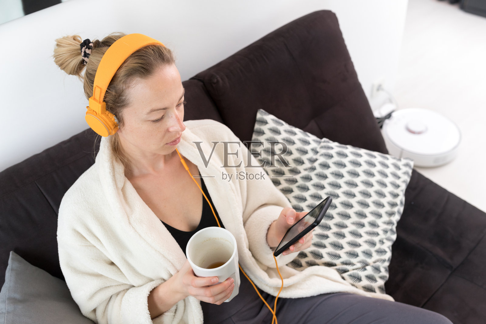 呆在家里。社会距离。女人坐在沙发上放松，喝着白色杯子里的茶，听着放松的音乐，通过手机上的社交网络与朋友和家人保持联系。照片摄影图片