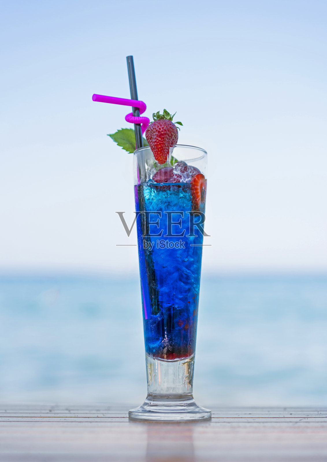 一杯新鲜的酒精蓝色鸡尾酒与烈酒，草莓片和冰块在日落的海滩上照片摄影图片