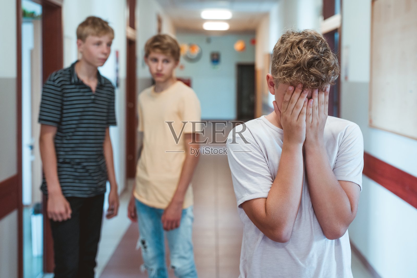 伤心受欺负的男孩站在学校走廊里，捂着脸。两个同学跟在他后面照片摄影图片