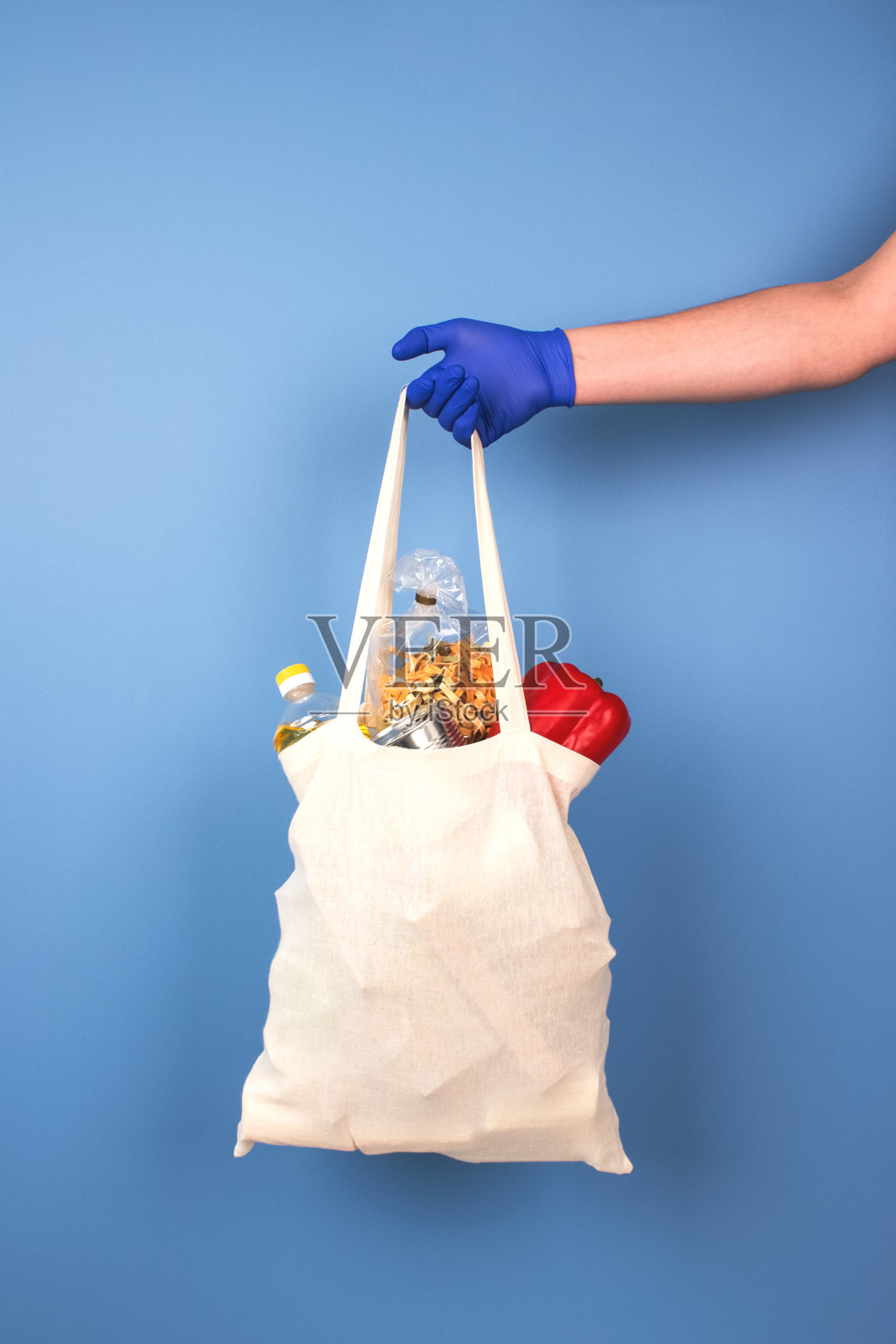 男人的手在蓝色一次性防护手套拿着白色袋子捐赠食物在蓝色时尚的背景。捐赠概念，模型和复制空间为您的设计，垂直构成照片摄影图片