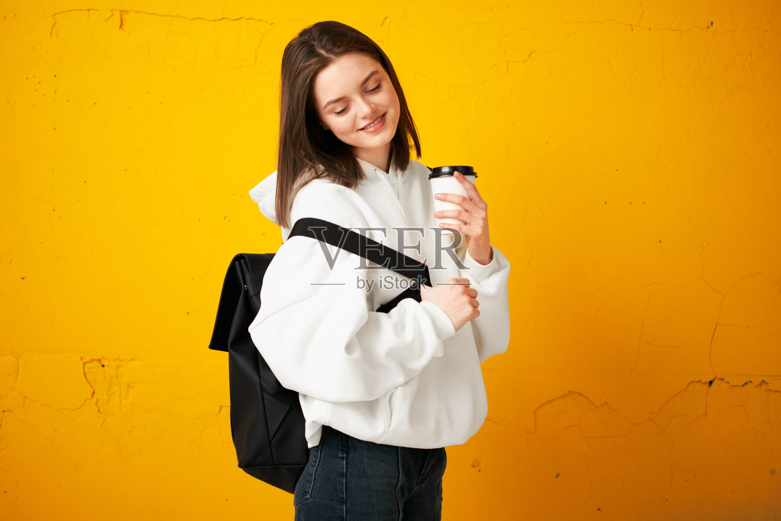 年轻漂亮的女孩穿着白色帽衫，背着背包，拿着一次性咖啡杯，靠在一堵黄色的混凝土墙上。照片摄影图片