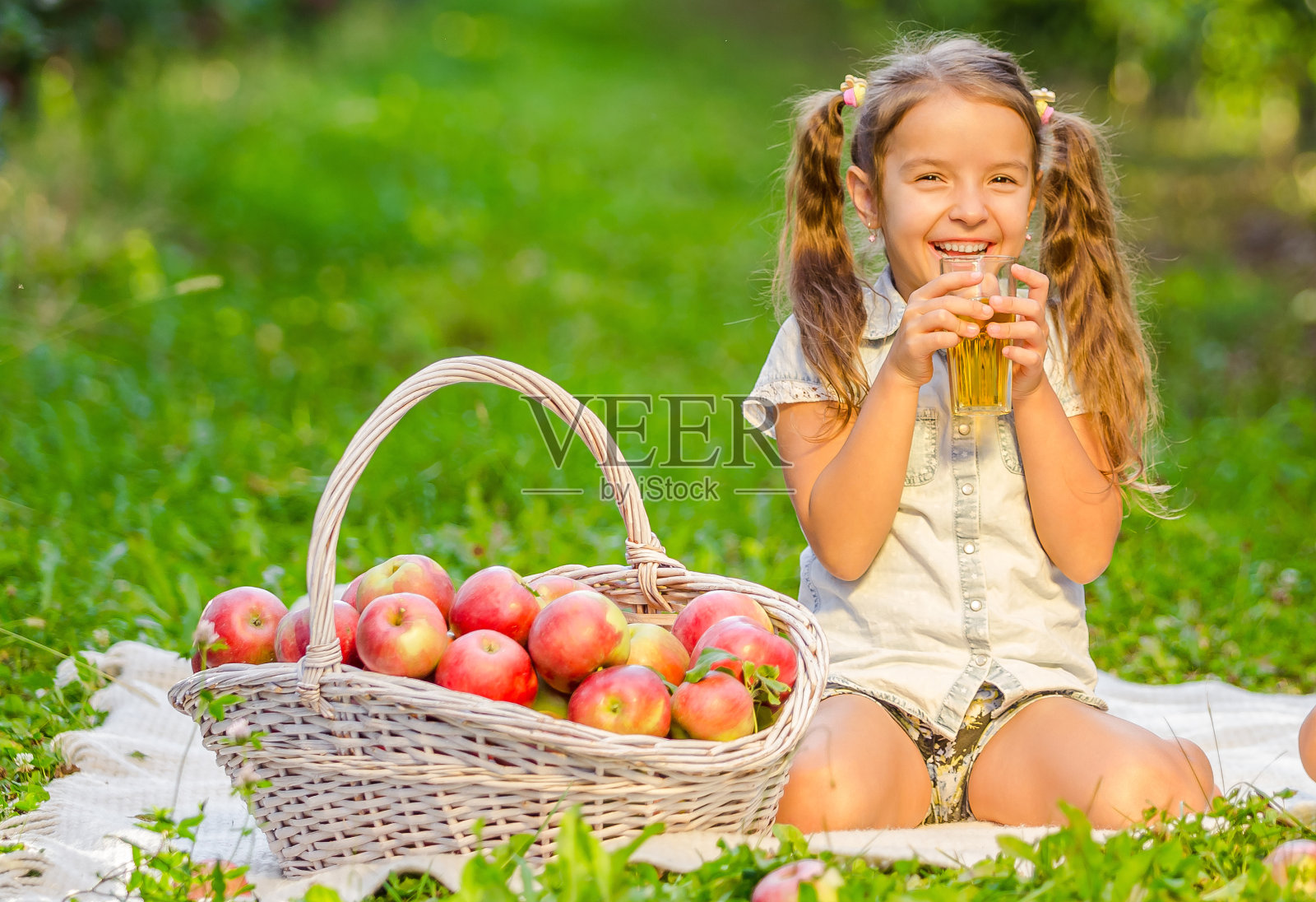 扎着马尾辫的小女孩在果园里喝着可口的苹果汁。照片摄影图片