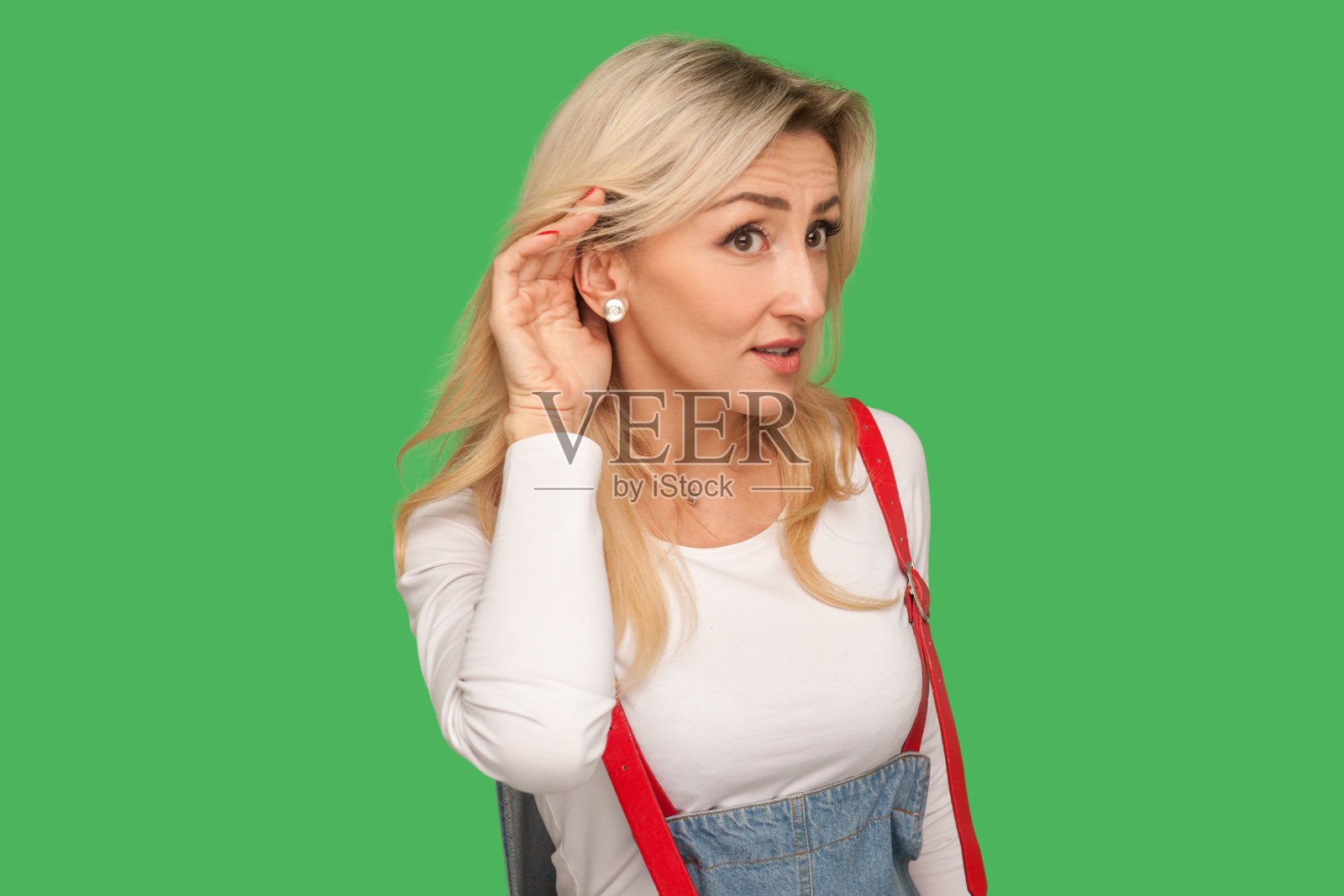 爱管闲事的好奇的成年金发女人在时尚的工装裤保持手靠近耳朵仔细听的肖像照片摄影图片
