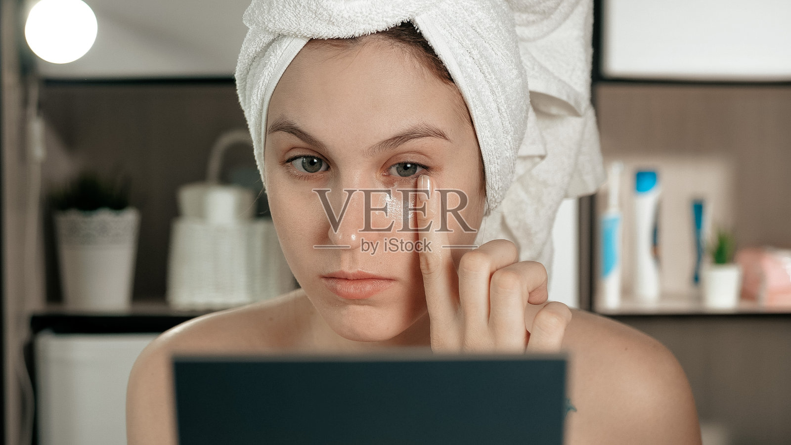 眼霜。年轻迷人的女人在她的左眼下涂抹护肤霜，女孩在浴室里看着镜子。卫生、美容、化妆、面部护理理念。特写镜头照片摄影图片