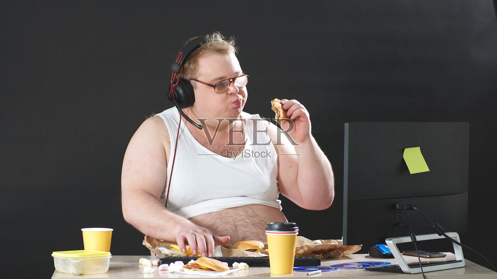 一个戴着眼镜和耳机坐在桌子前一边吃一边玩电脑游戏的胖子。闭关锁国则,摧毁检疫照片摄影图片