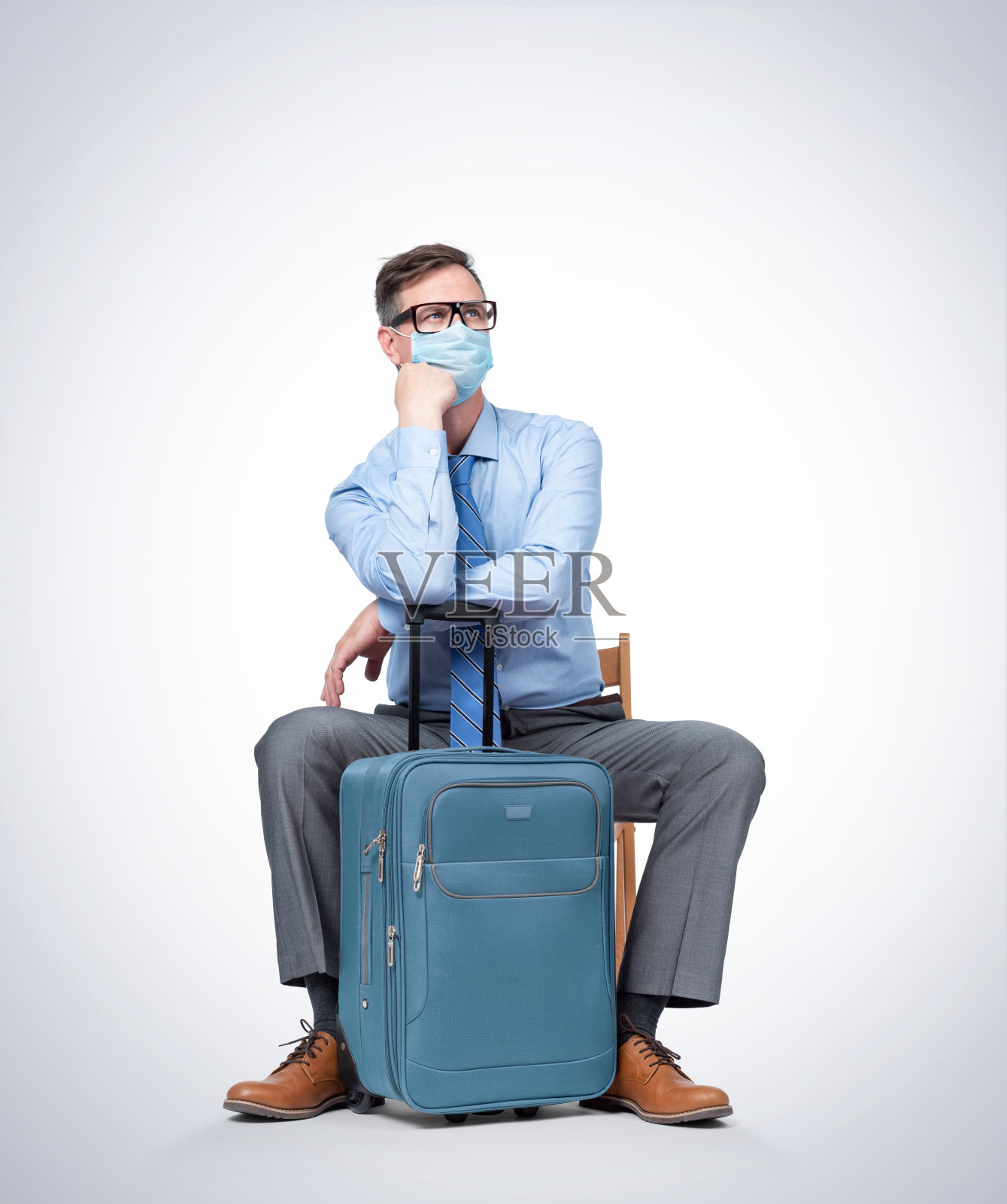 一个戴着呼吸面罩、眼镜、衬衫和领带的男人坐在一个手提箱旁等待飞机。流行的旅行概念。照片摄影图片