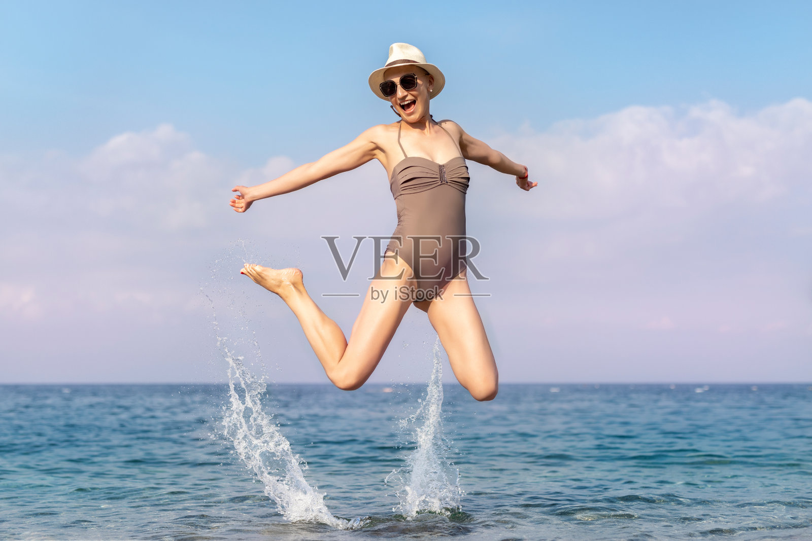 年轻的成年人秃顶剃发时髦漂亮的白人女性在泳装帽子和太阳眼镜跳跃和跳跃在海洋或海洋海岸海滩避暑胜地。青春健康周末快乐理念照片摄影图片