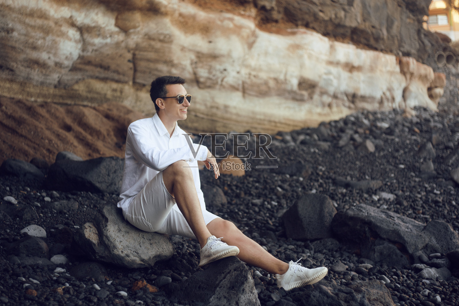一个人坐在海洋的岩石上。特内里费岛的石滩。一个穿着白色衣服，戴着眼镜的男人望着远方。一个男人正在海滩上放松，沉思，平静，沉思。照片摄影图片