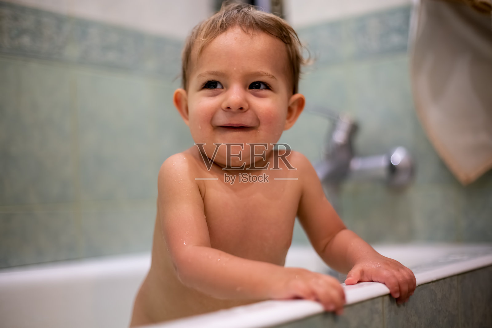幸福的概念。快乐可爱的小宝贝站在家里洗澡微笑。背景是一间模糊的绿色浴室。特写,软焦点照片摄影图片