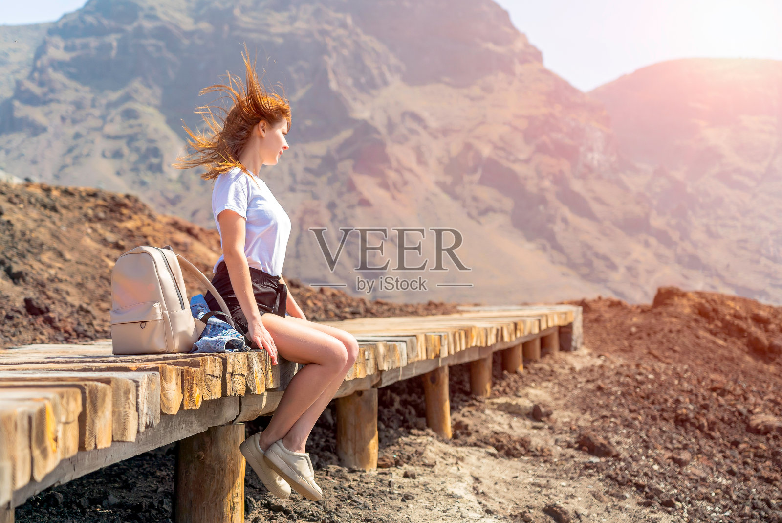 一个背着背包的年轻女子坐在岩石海岸的木制平台上。快乐的女孩在一个晴朗的日子与明亮的蓝色天空。背景是特内里费岛的火山。照片摄影图片