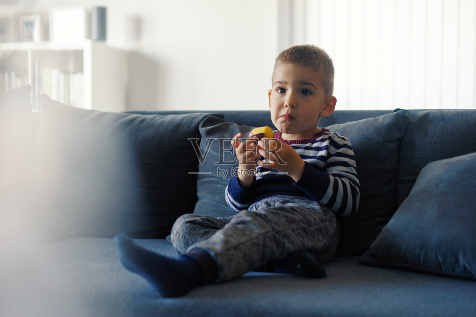 一个三岁的白人小男孩坐在家里的沙发床上，手里拿着巧克力糖，一边吃一边看电视照片摄影图片