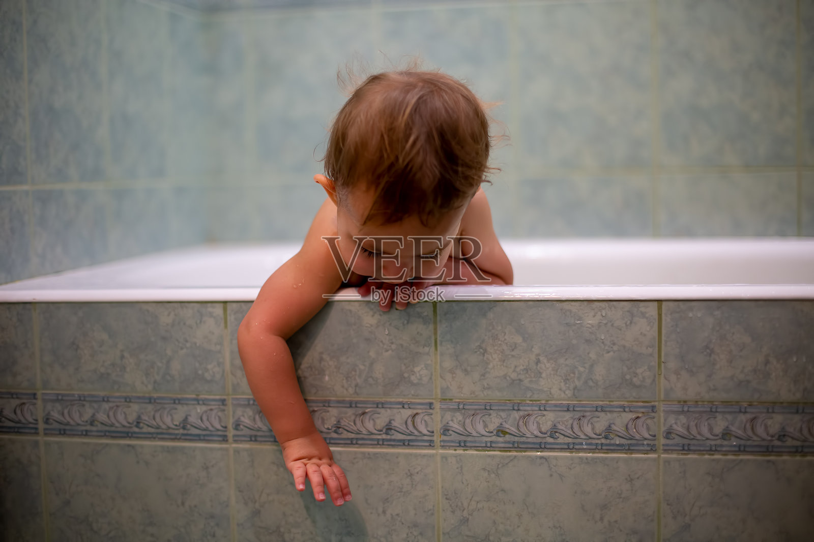 一个可爱的小宝贝，洗澡的时候，从浴缸的一边挂下来，一只手伸展下来。以模糊的绿色浴室为背景照片摄影图片