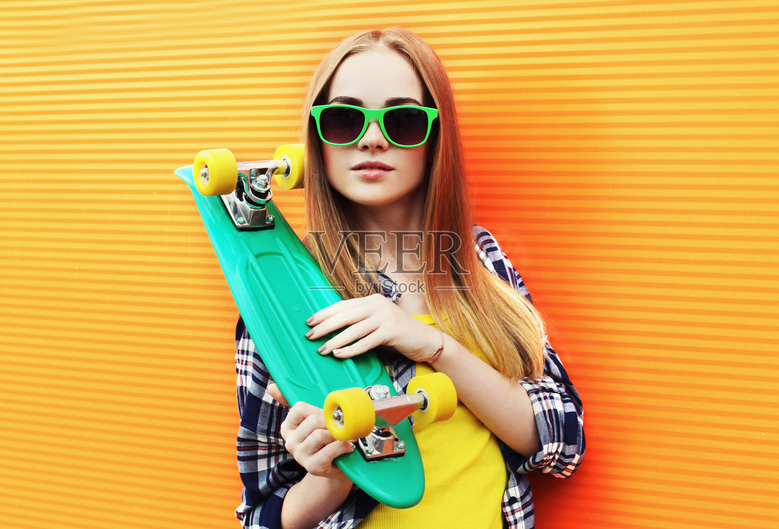 肖像年轻的金发女子与绿色滑板戴太阳镜在彩色的橙色墙背景照片摄影图片