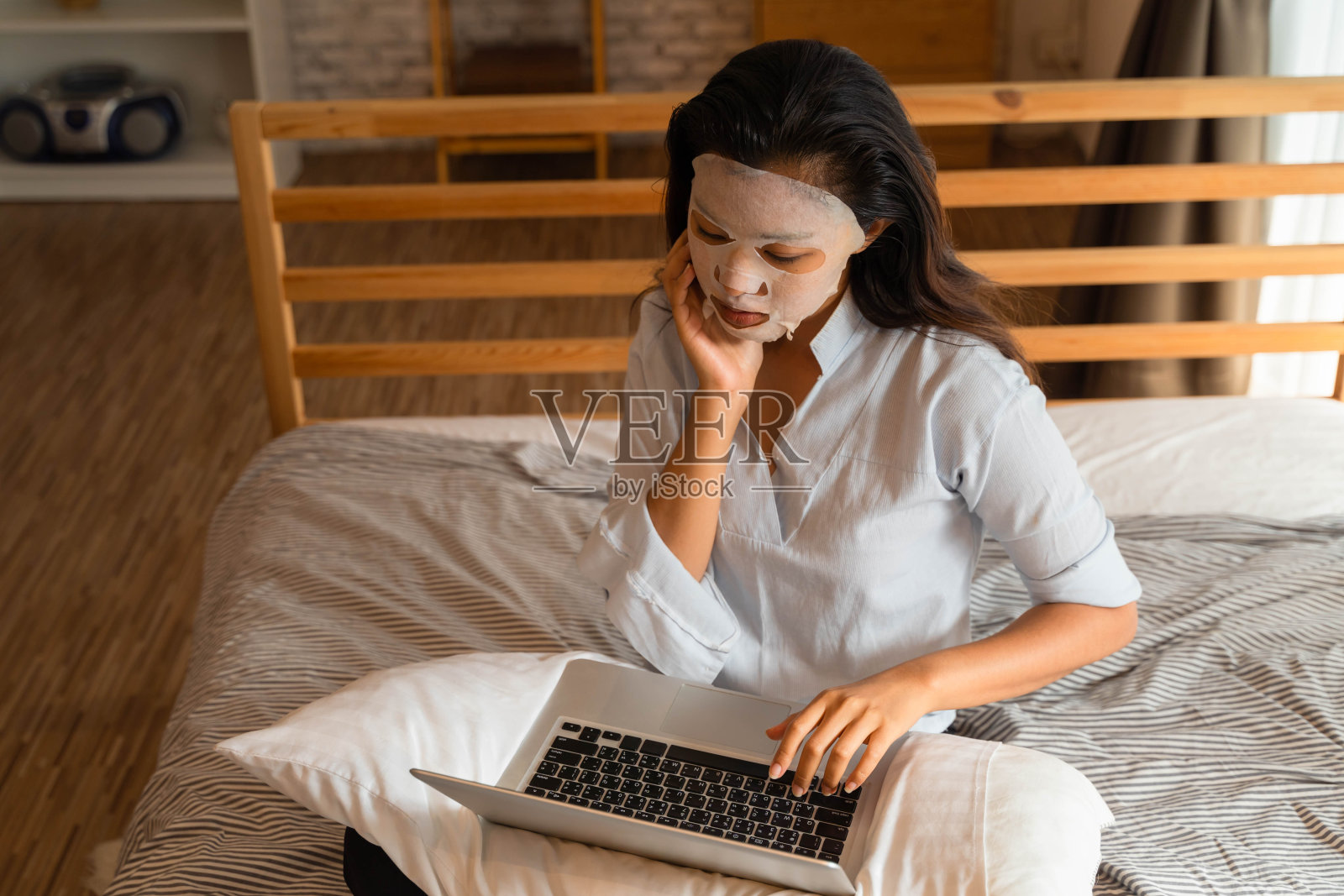 一名年轻的亚洲女孩在卧室里用笔记本电脑时敷面膜的肖像。女性美容和面部护理概念照片摄影图片