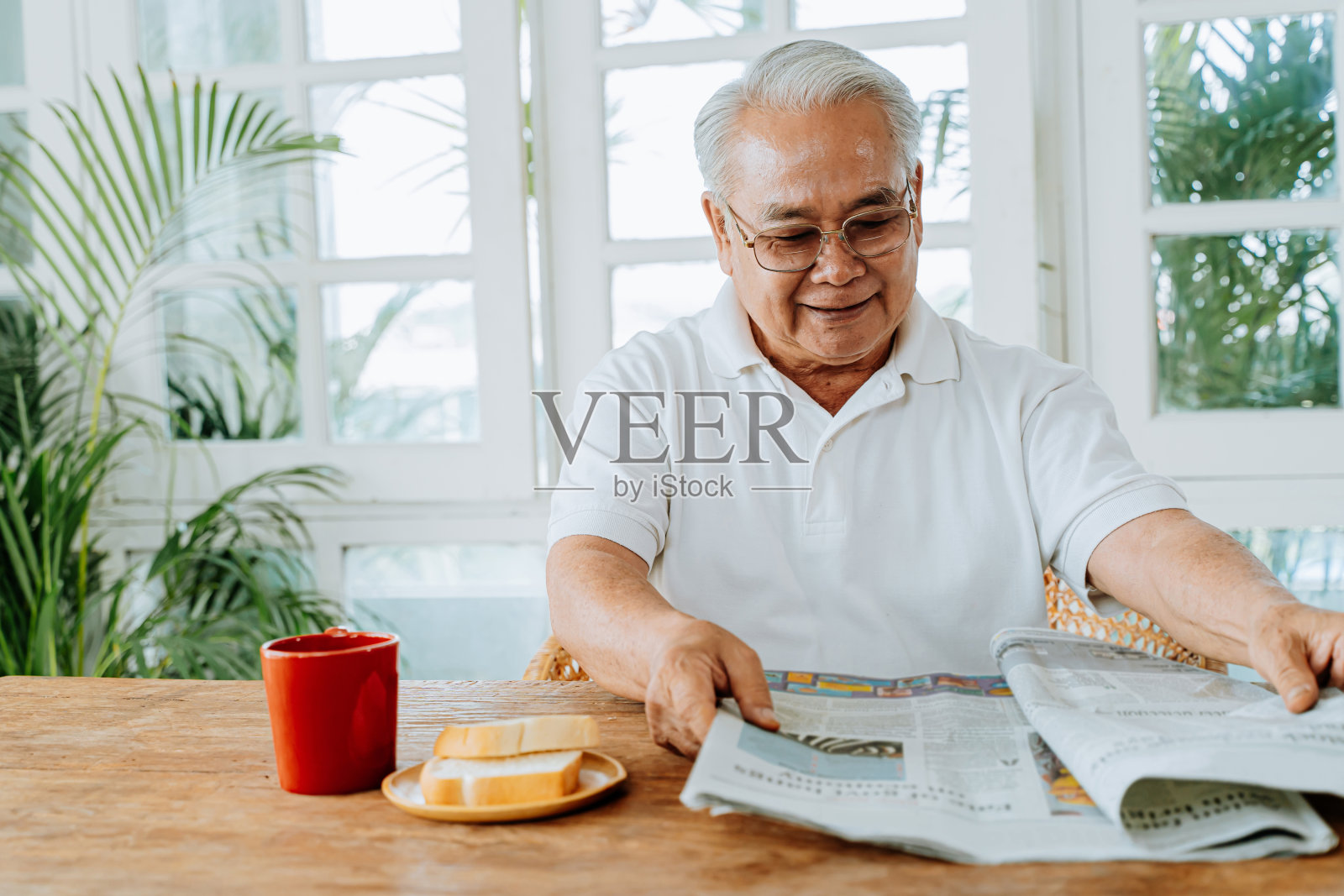 快乐的资深亚洲男性在休闲衣服和眼镜阅读有趣的文章，在家里吃早餐。享受退休生活的老人照片摄影图片
