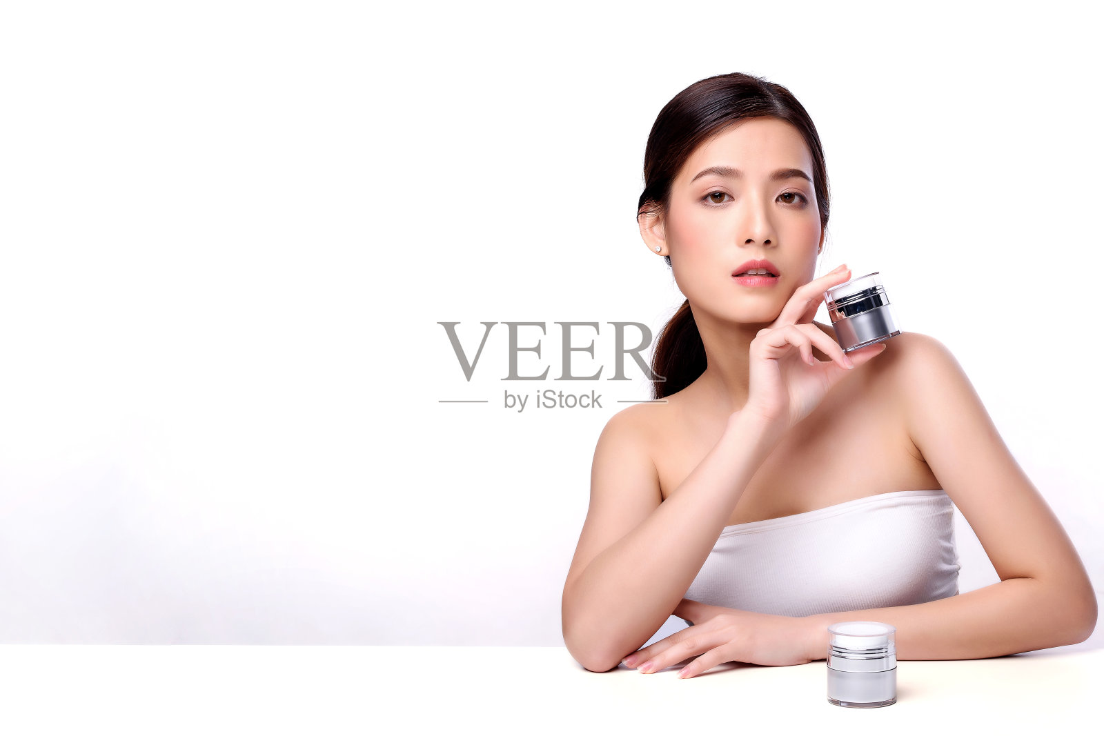肖像美丽迷人的年轻亚洲女人摆面部化妆品和坐在白色的桌子，美丽的女性脸与皮肤护理在白色的背景，保健和皮肤护理的概念照片摄影图片