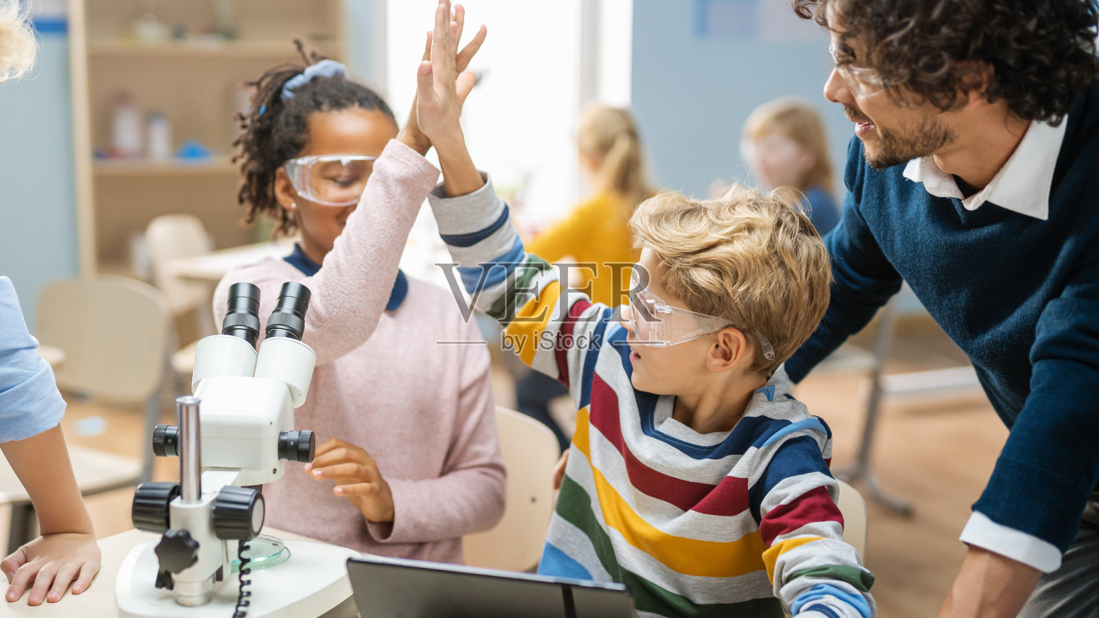 小学科学教室:可爱的小女孩用显微镜看，男孩用数码平板电脑在网上查信息，他们庆祝击掌。照片摄影图片