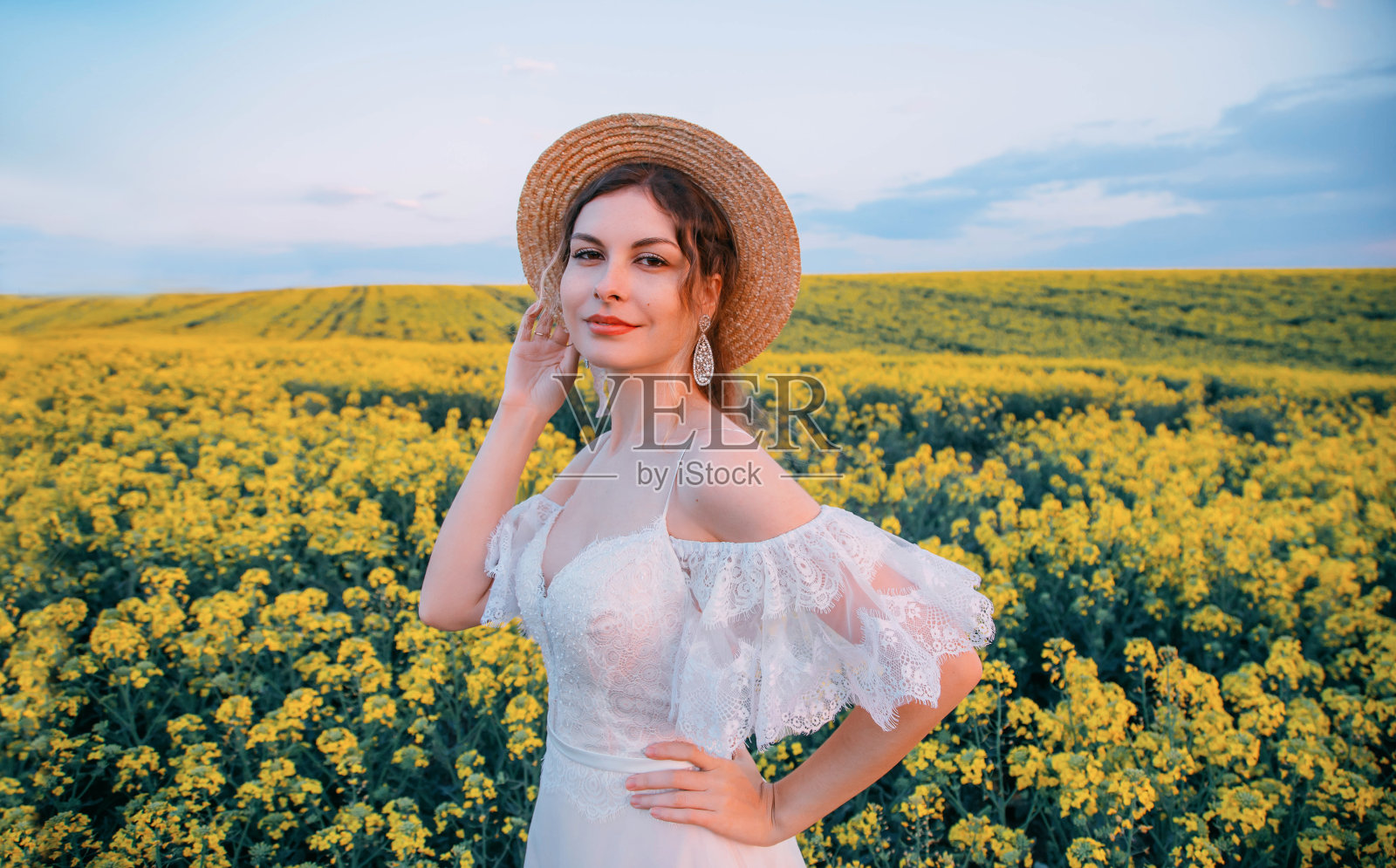 描绘年轻快乐的乡村复古女人。稻草的帽子。奢华的白色婚纱。背景是春花盛开的油菜籽田。明亮的夏天的天空。自然的妆容。可爱的笑脸照片摄影图片
