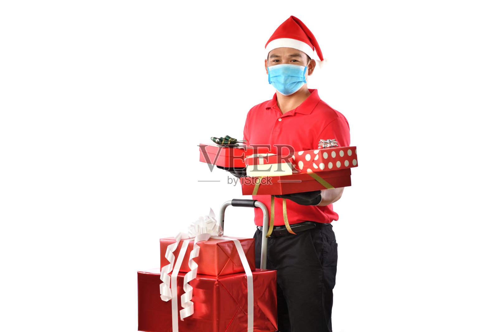 在新冠肺炎疫情爆发和圣诞节期间，身穿红色制服、佩戴医用口罩、防护手套、戴圣诞帽的快乐年轻亚洲快递员手持装有礼物的盒子，双手被隔离在白色背景上照片摄影图片