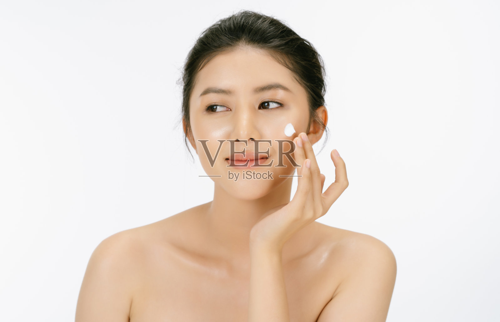 美丽的年轻亚洲女人裸露的肩膀是应用面霜在她的脸上和和看远处孤立的白色背景。女性美脸护肤理念。照片摄影图片