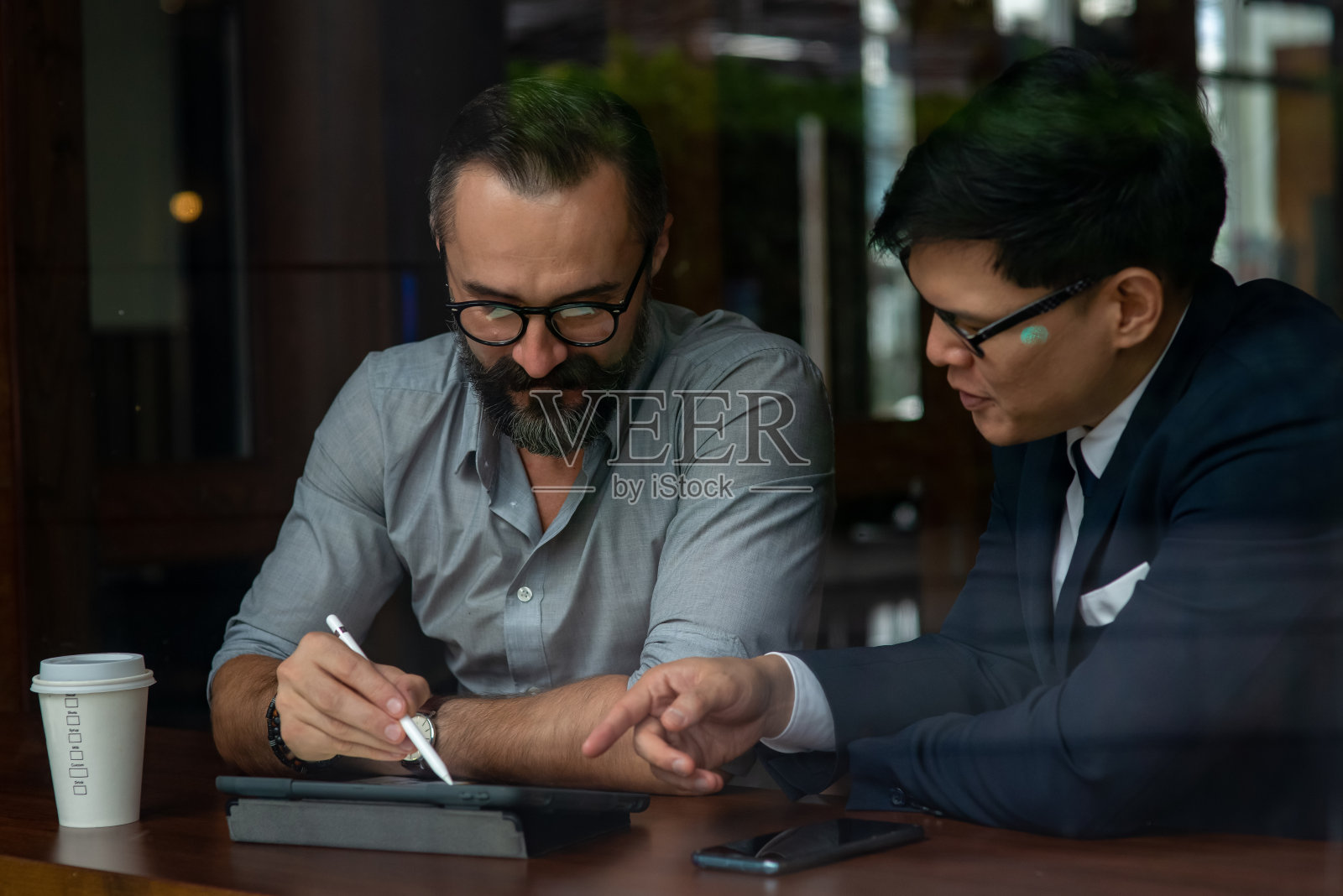 白种人胡子男子创意自由职业者坐在咖啡店与亚洲商人在西装讨论商业计划与使用数字平板电脑。商务会议团队合作和技术理念。照片摄影图片