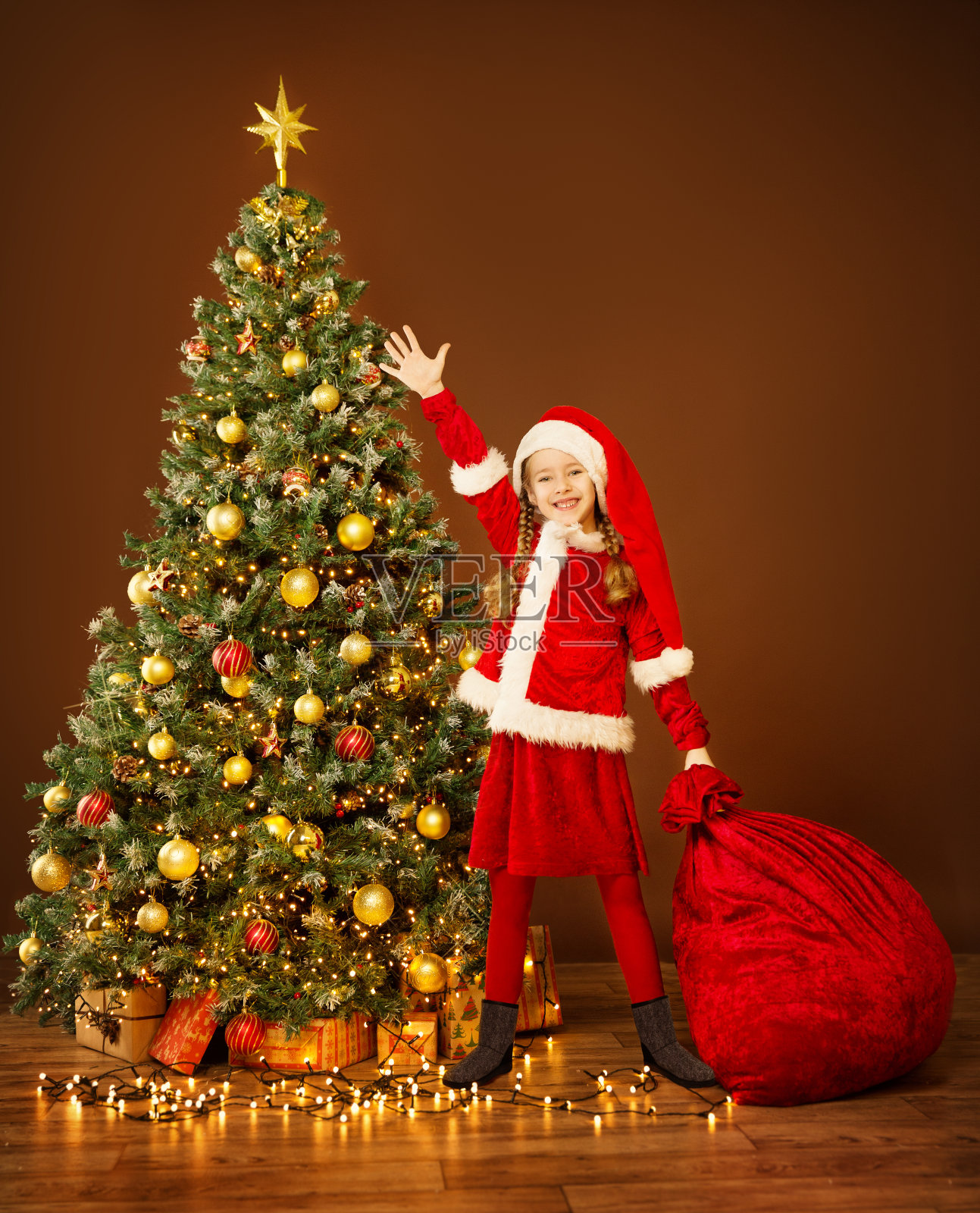 圣诞快乐的孩子在精灵服装拿着红色圣诞包新年礼物在圣诞树下。装饰照明家居照片摄影图片