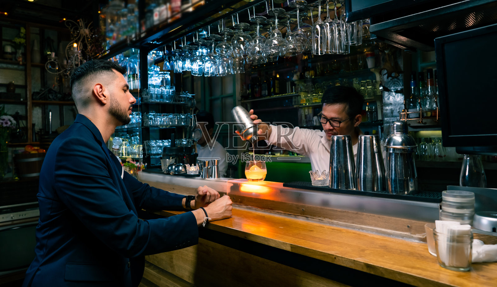肖像英俊的白人男子坐在吧台和等待美味的鸡尾酒从酒吧的酒吧招待在夜总会。亚洲男性调酒师调酒师准备在岩石玻璃上为顾客混合酒精饮料照片摄影图片
