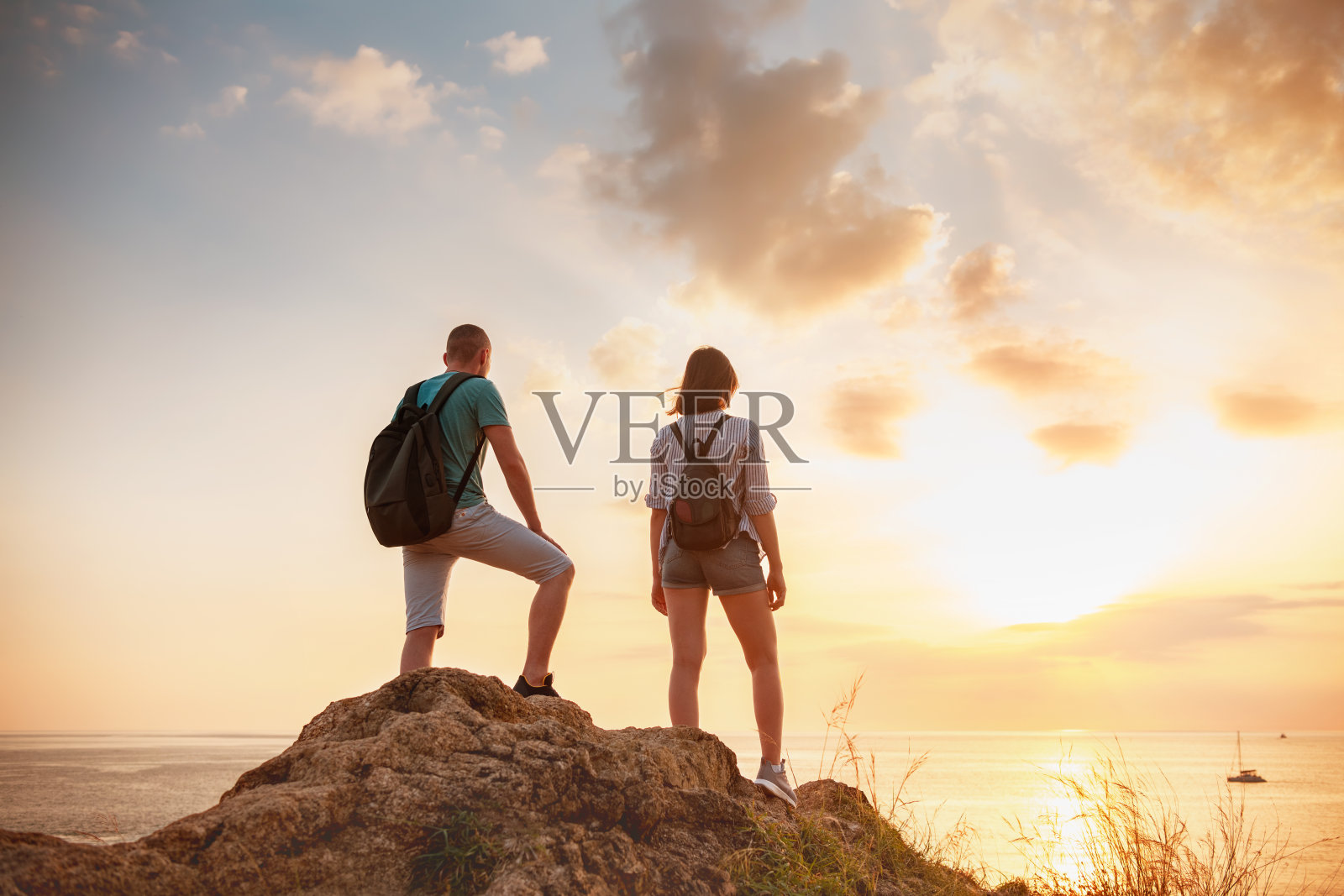 两个徒步旅行者站在一块大岩石上，看着夕阳下的大海照片摄影图片