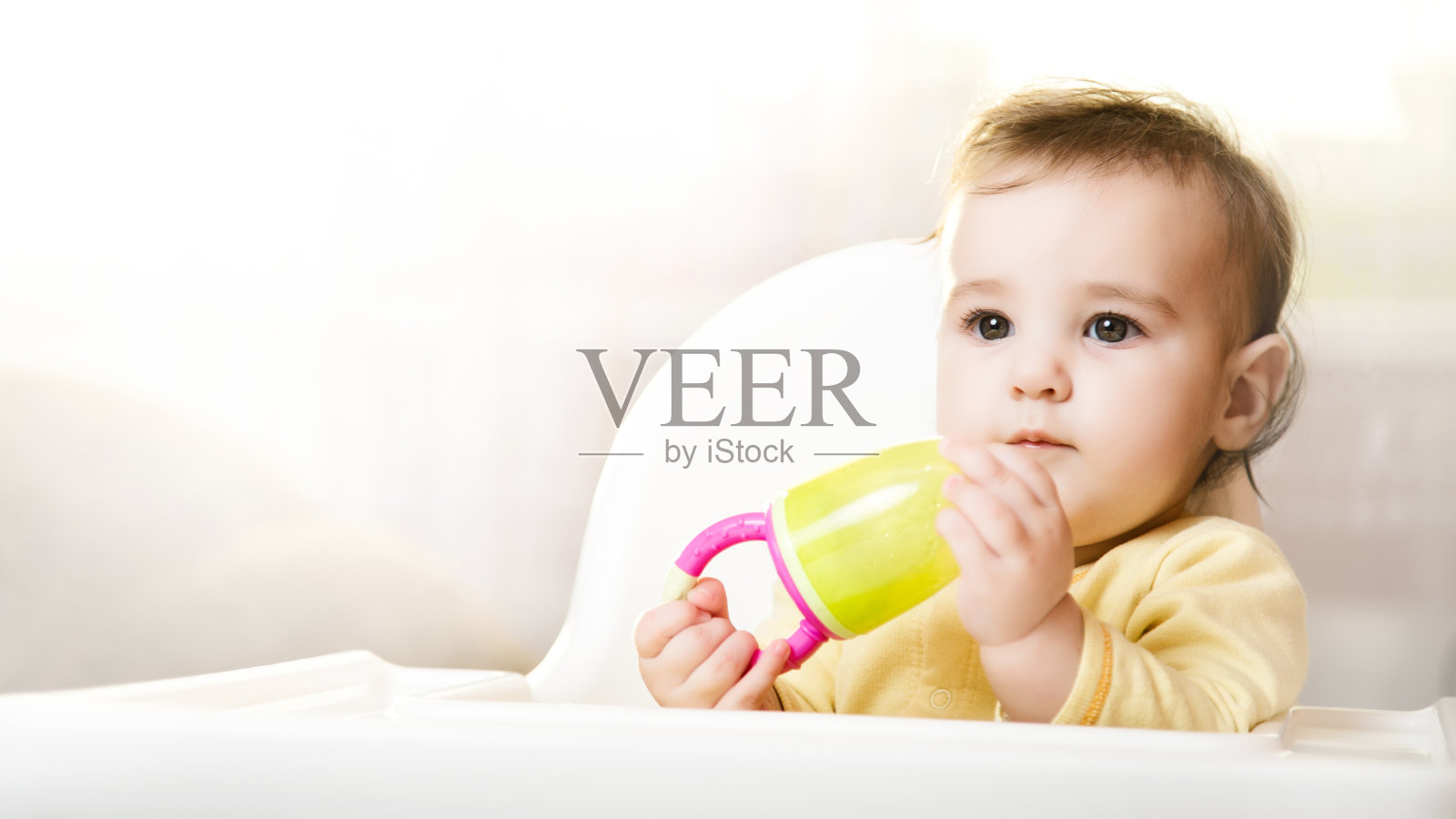 小婴儿坐在高椅子上，在室内吃食物。婴儿的喂养。6个月大的蹒跚学步的孩子，手里拿着小嚼器照片摄影图片