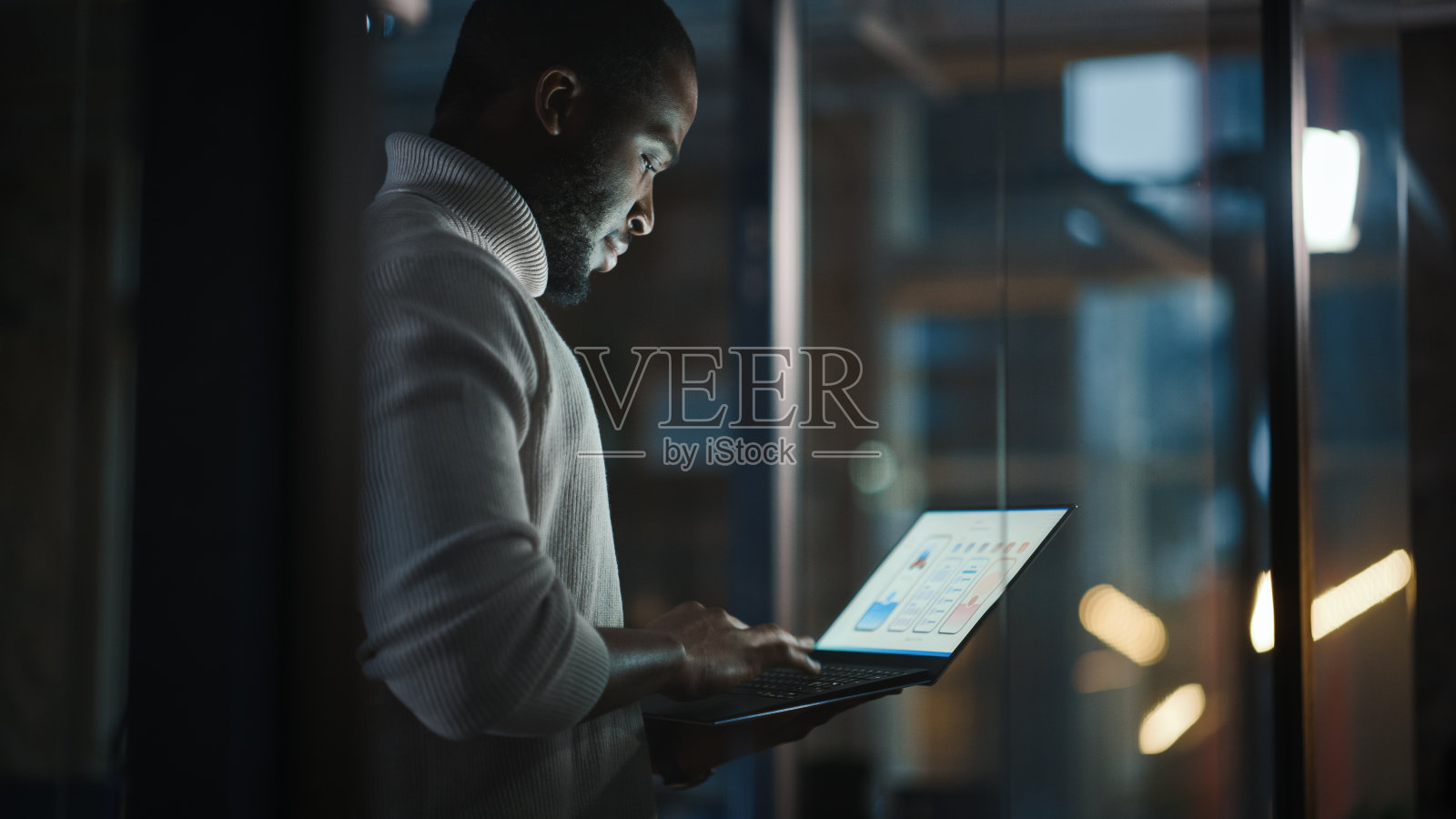 一位英俊的非裔美国黑人男性正站在一家创意公司的会议室里，手里拿着笔记本电脑。身穿白色毛衣的项目经理，致力于App用户界面的开发。照片摄影图片