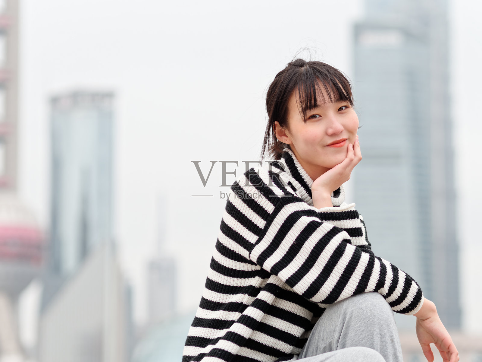 冬日里，一名中国年轻女子手捂着脸对着镜头微笑，背景是上海的地标建筑。照片摄影图片