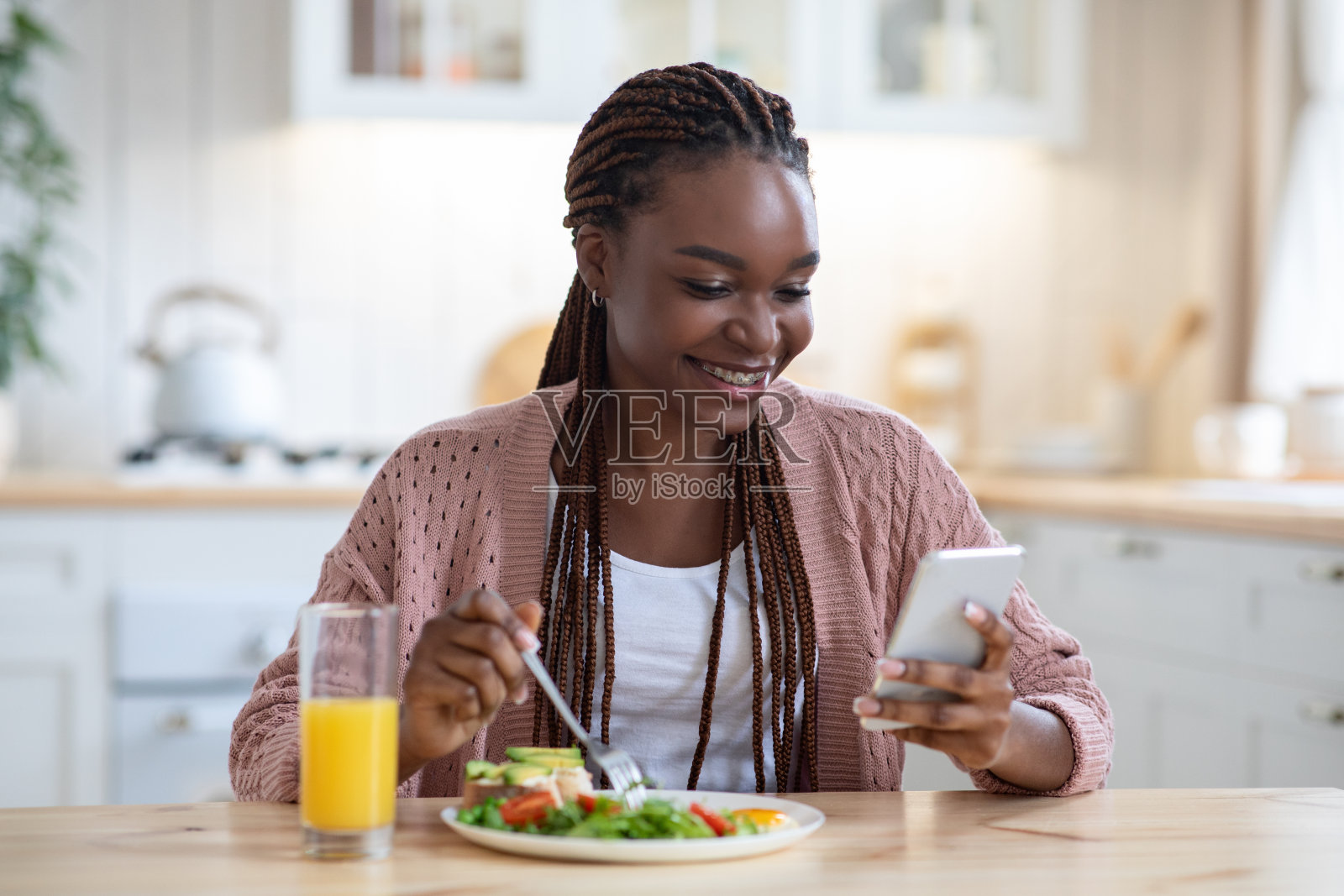 微笑的黑人妇女在厨房用智能手机吃早餐照片摄影图片