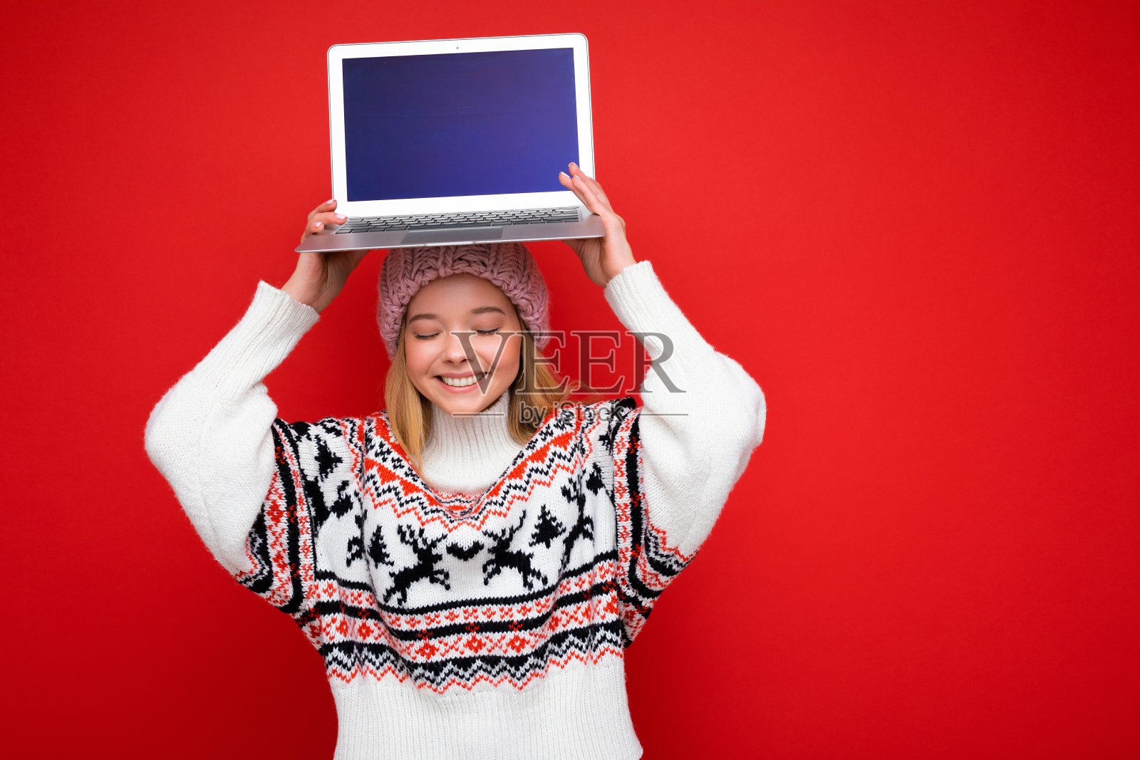 照片拍摄的美丽微笑的金发年轻女子拿着电脑笔记本电脑与空白的显示器屏幕模拟和复制空间穿着针织冬季帽子和毛衣闭上眼睛和微笑的嘴孤立在红色墙的背景照片摄影图片