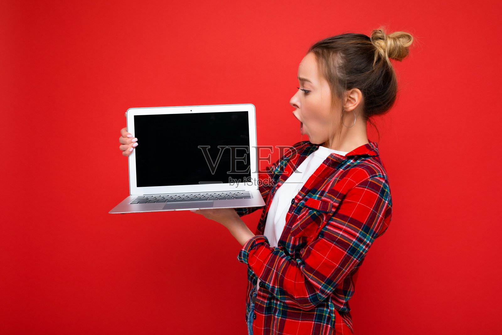 美丽的震惊的年轻女子拿着笔记本电脑，穿着红色的衬衫看着隔离在红色背景上的显示器照片摄影图片