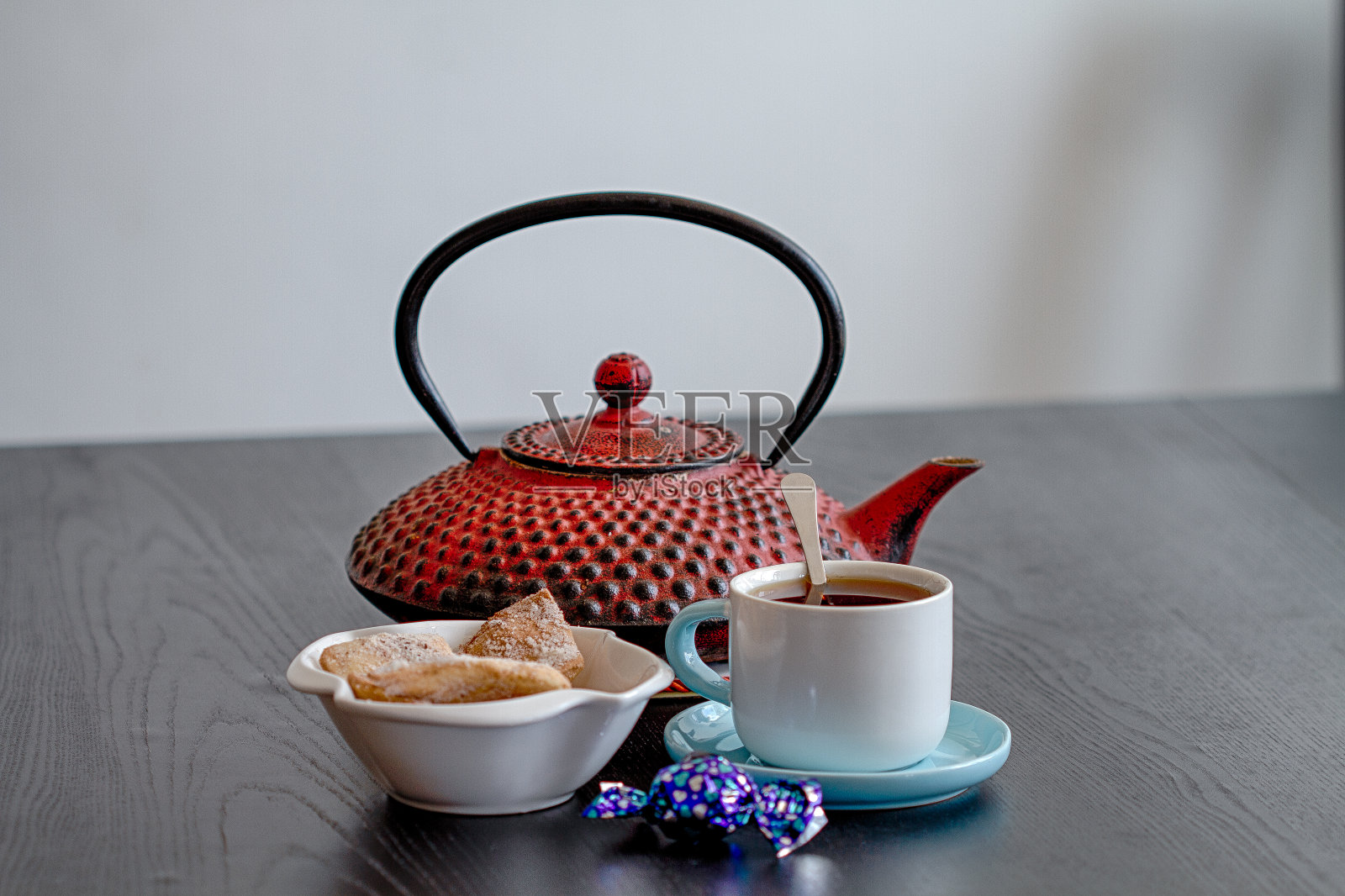 黑色的桌子上放着一只红色茶壶，背景上放着一杯带饼干的茶。照片摄影图片