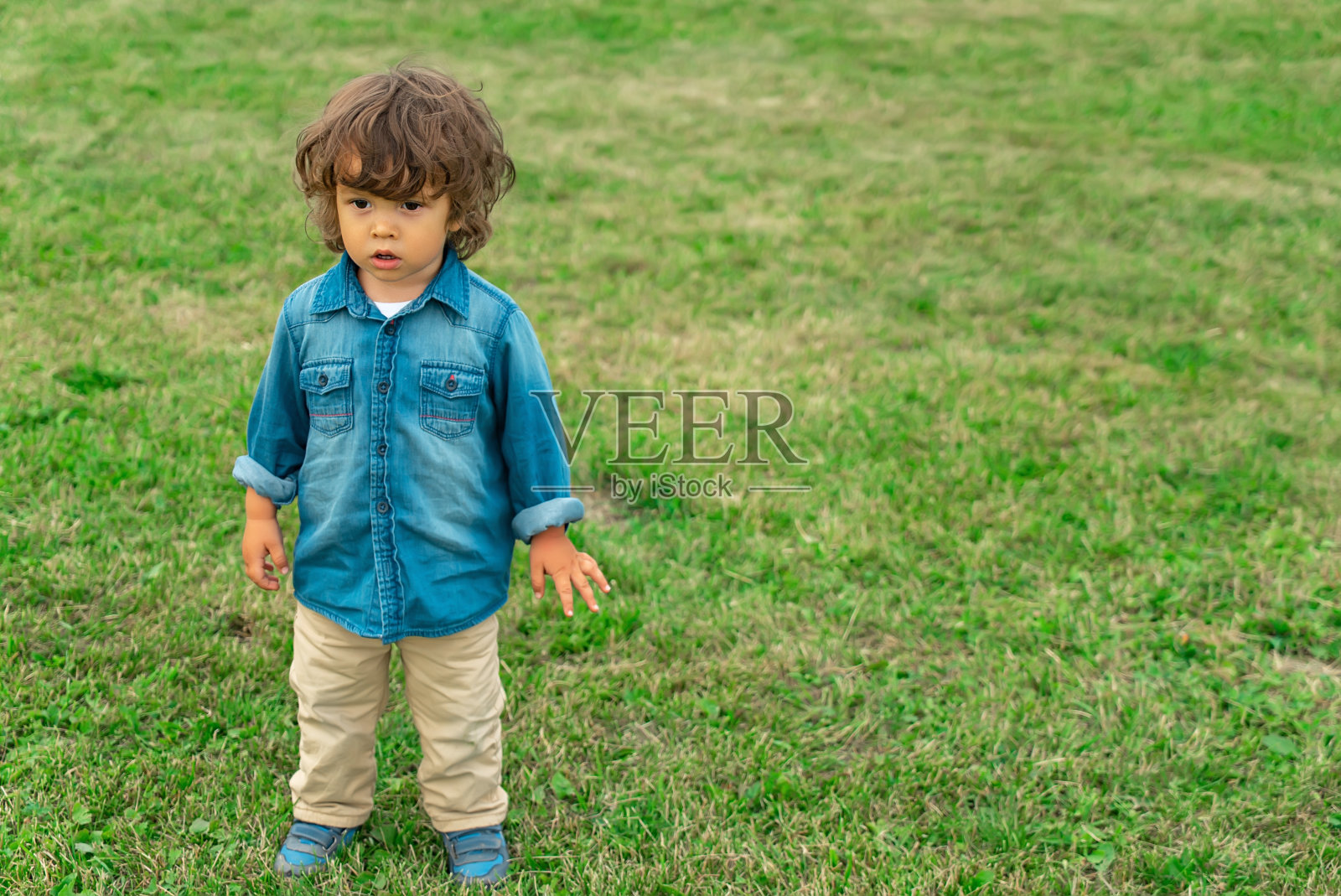长头发可爱的小男孩站在户外的草坪上。照片摄影图片