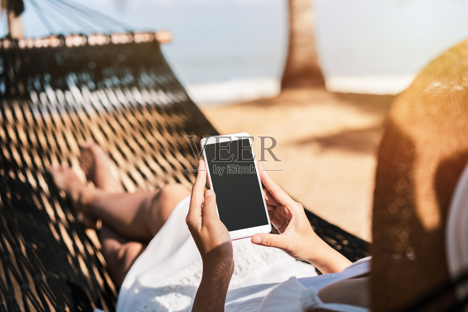一名年轻女子在暑假旅行时，躺在海滩上的吊床上使用智能手机照片摄影图片