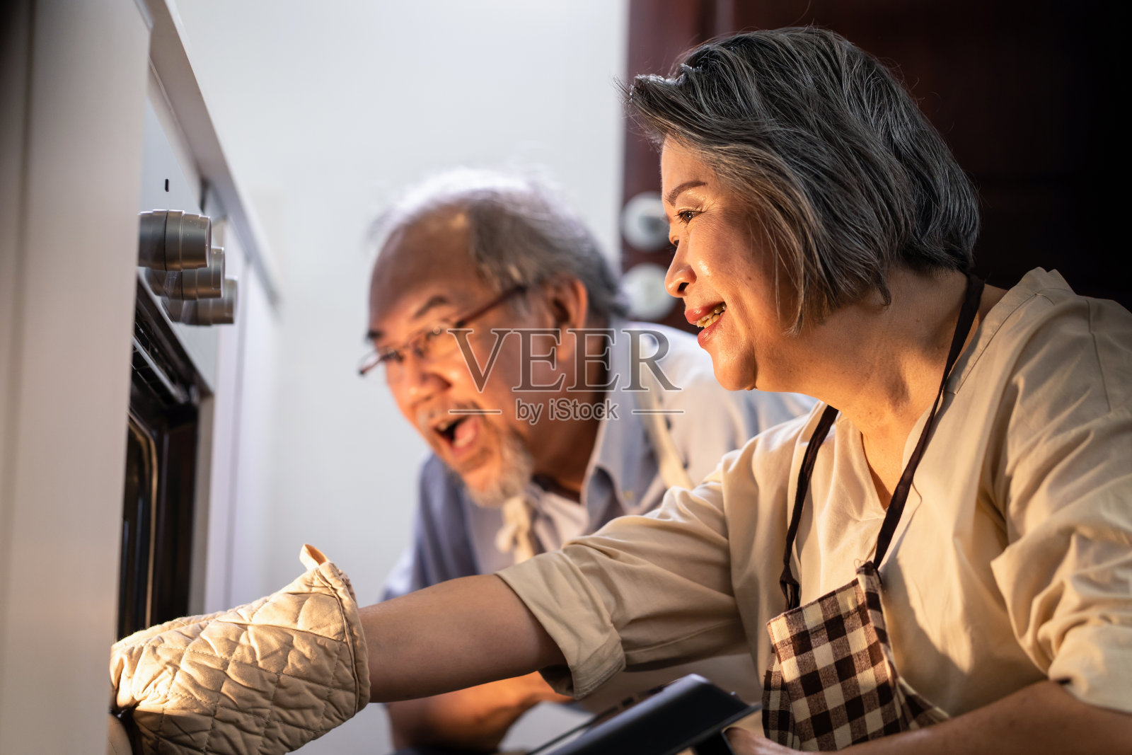 亚洲老人在家里做披萨。一位老妇人打开烤箱，把食物从机器里拿出来。老人看着饭，微笑着闻着，一起享受家庭活动。照片摄影图片