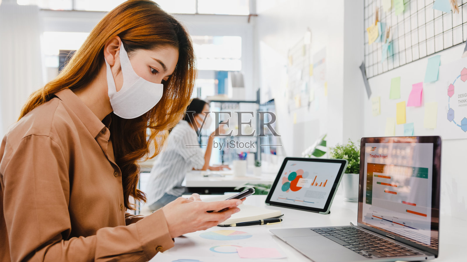 亚洲女企业家在新常态下戴着口罩进行社交距离以预防病毒，同时在办公室使用笔记本电脑和电话。照片摄影图片