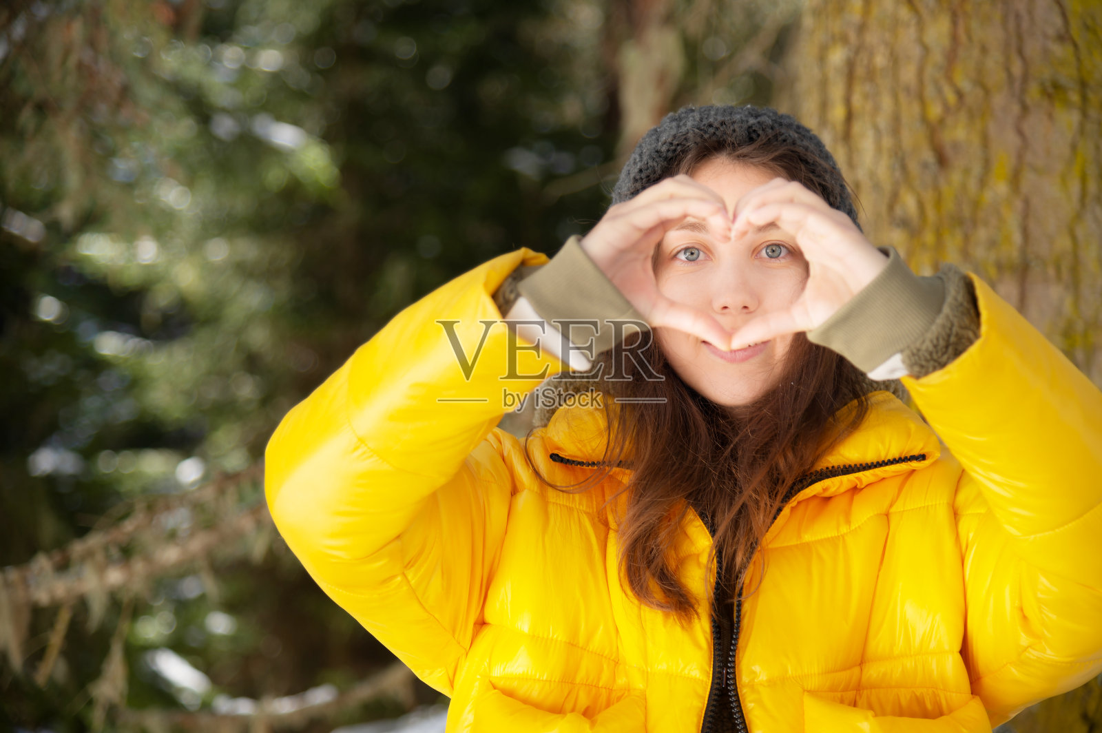 迷人的年轻白种人女子在羽绒服和帽子显示一种姿态的心和爱的相机与她的手。背景是冬季的针叶林照片摄影图片